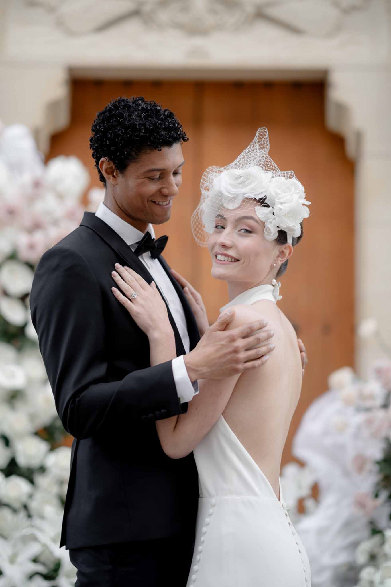 Couple embracing before arched door with birdcage veil and white and blush rose arrangements