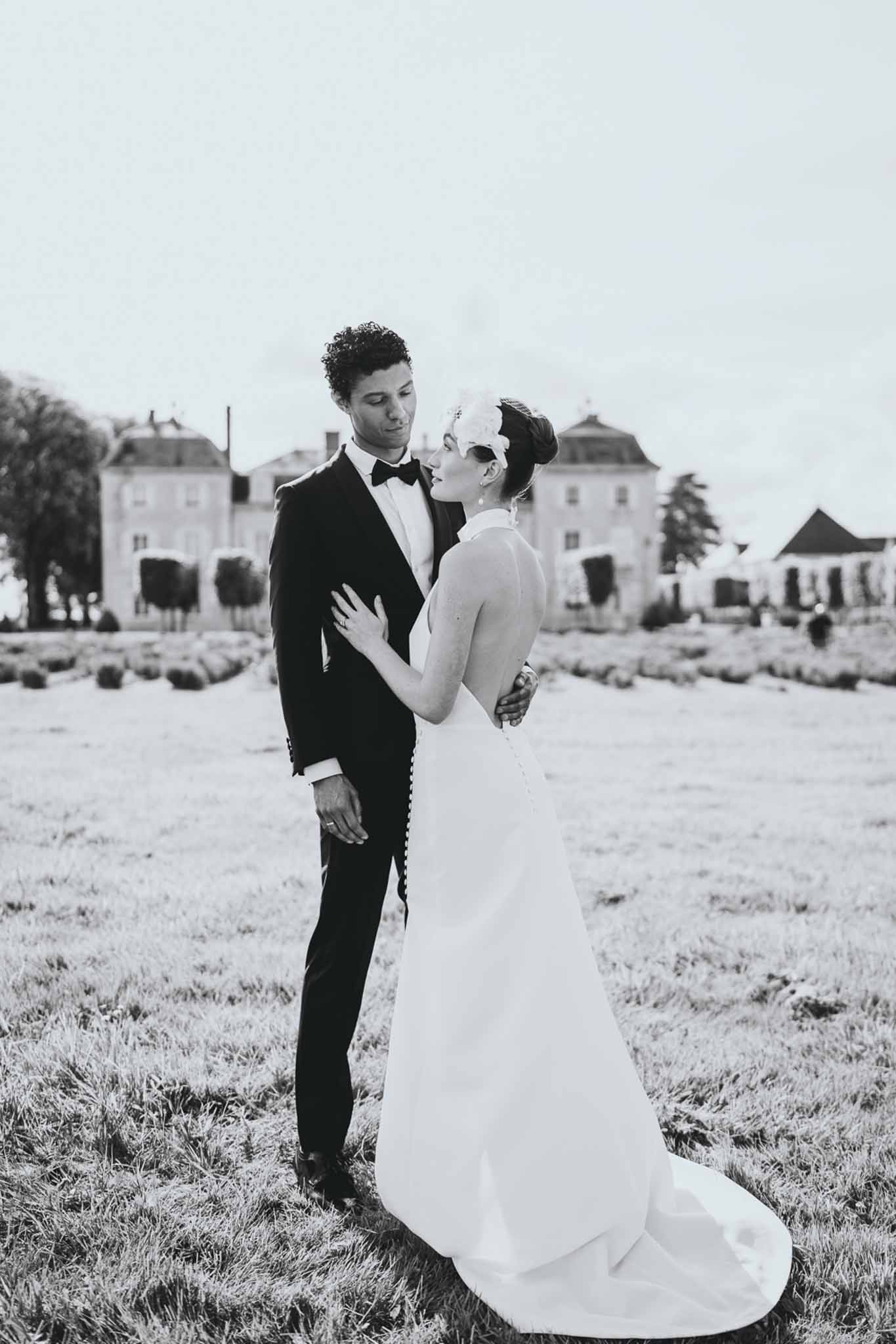 Black and white portrait of bride and groom facing each other on chateau grounds with manor house behind