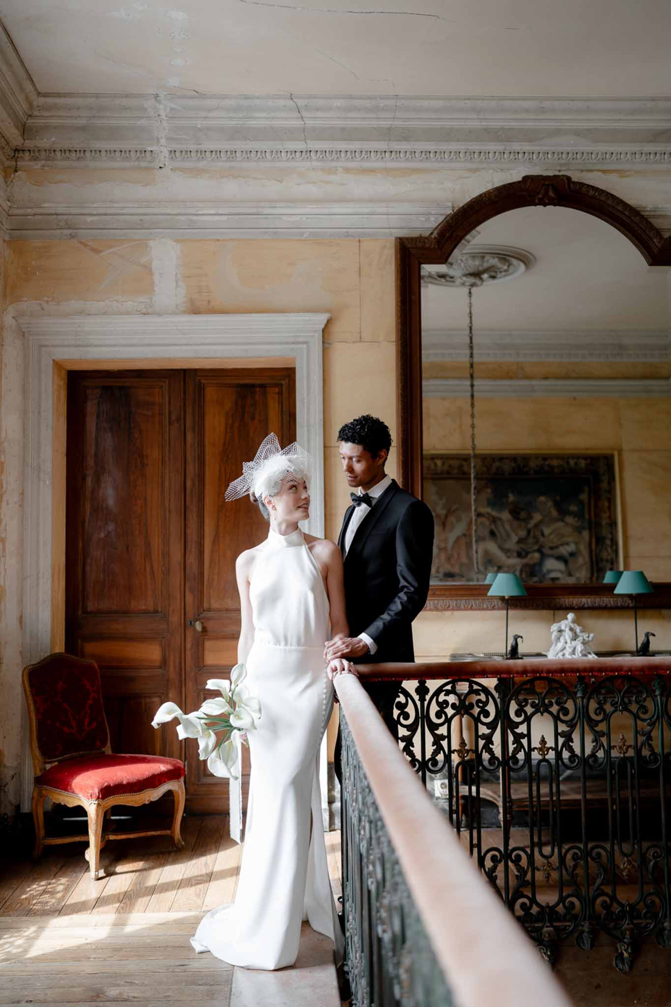 Bride in ivory halter gown with birdcage fascinator and calla lily bouquet beside groom on chateau mezzanine landing