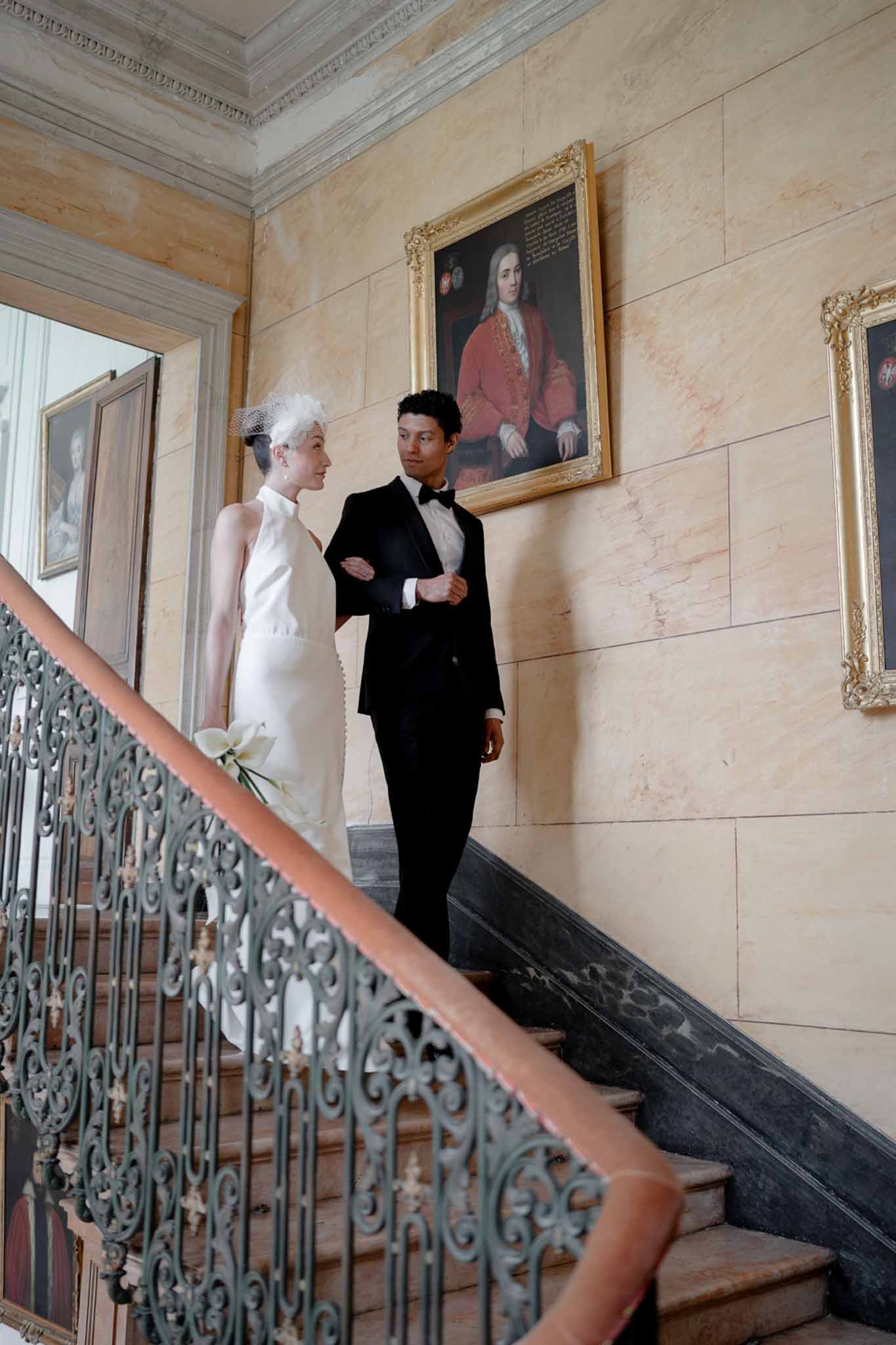 Bride in halter gown with birdcage veil and groom in tuxedo on marble staircase with gilt portraits