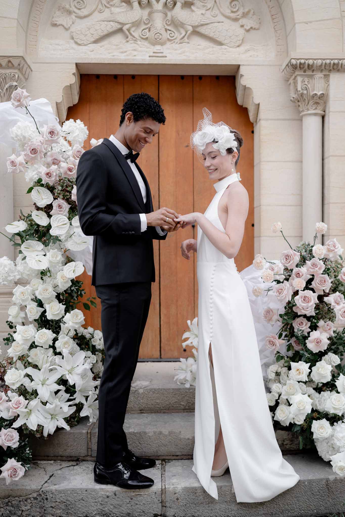 Groom placing ring on bride's finger flanked by white rose and lily floral installations on stone steps