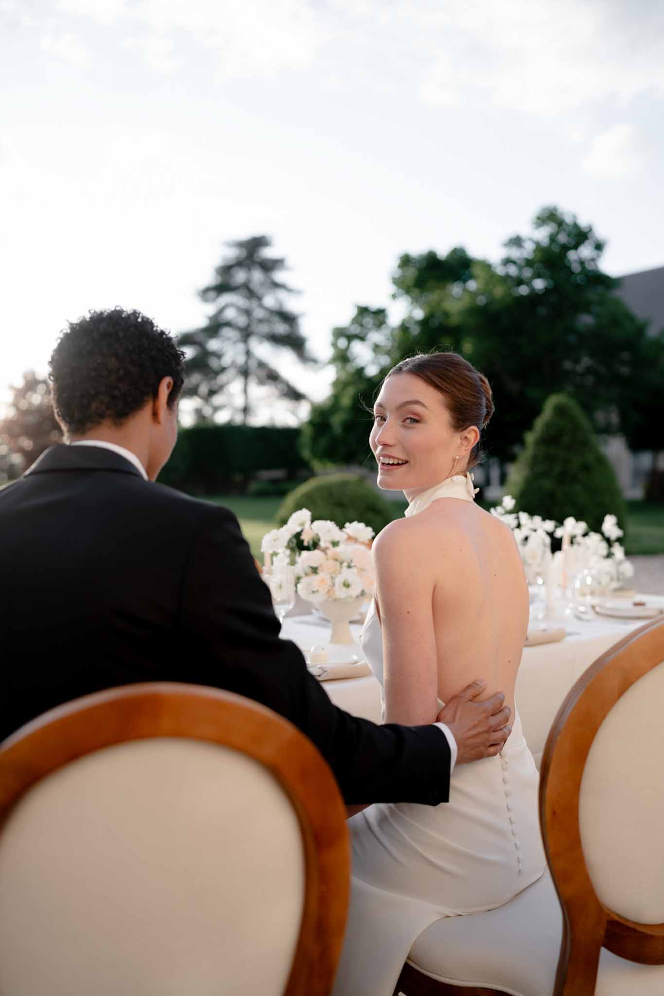 Bride in backless ivory halter gown and groom at reception table with blush floral centerpieces and oval-back chairs