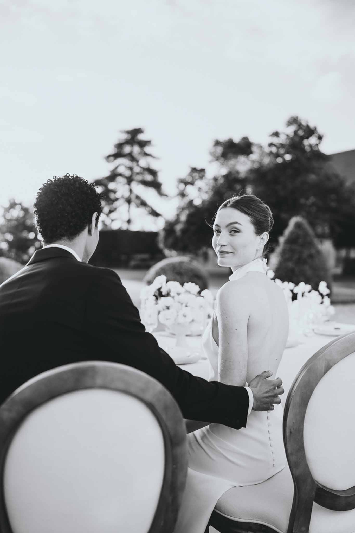 Black and white photo of bride and groom seated at reception table showing back of halter gown with button details