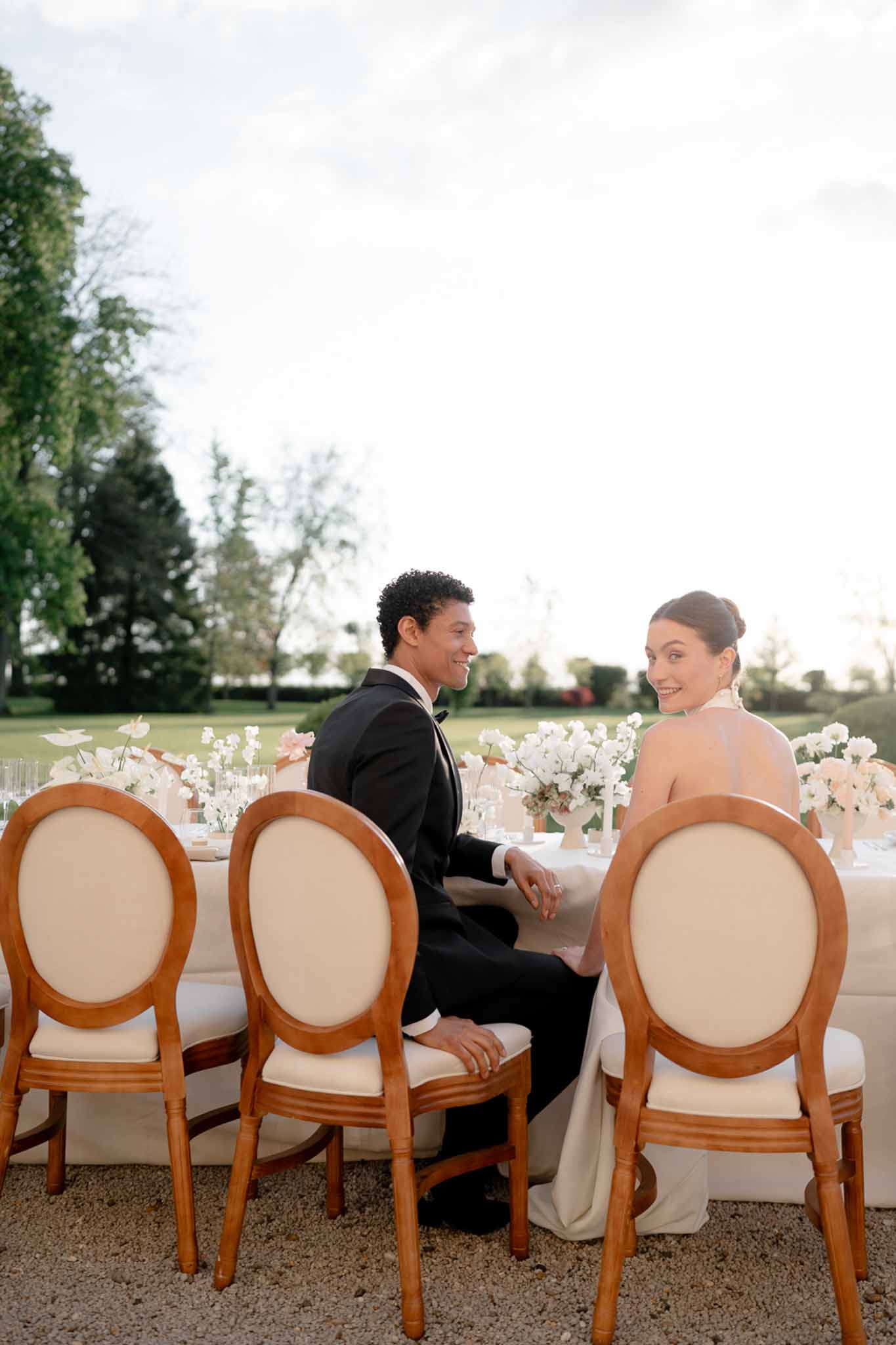 Bride and groom smiling at each other at an outdoor reception table with white and blush floral arrangements