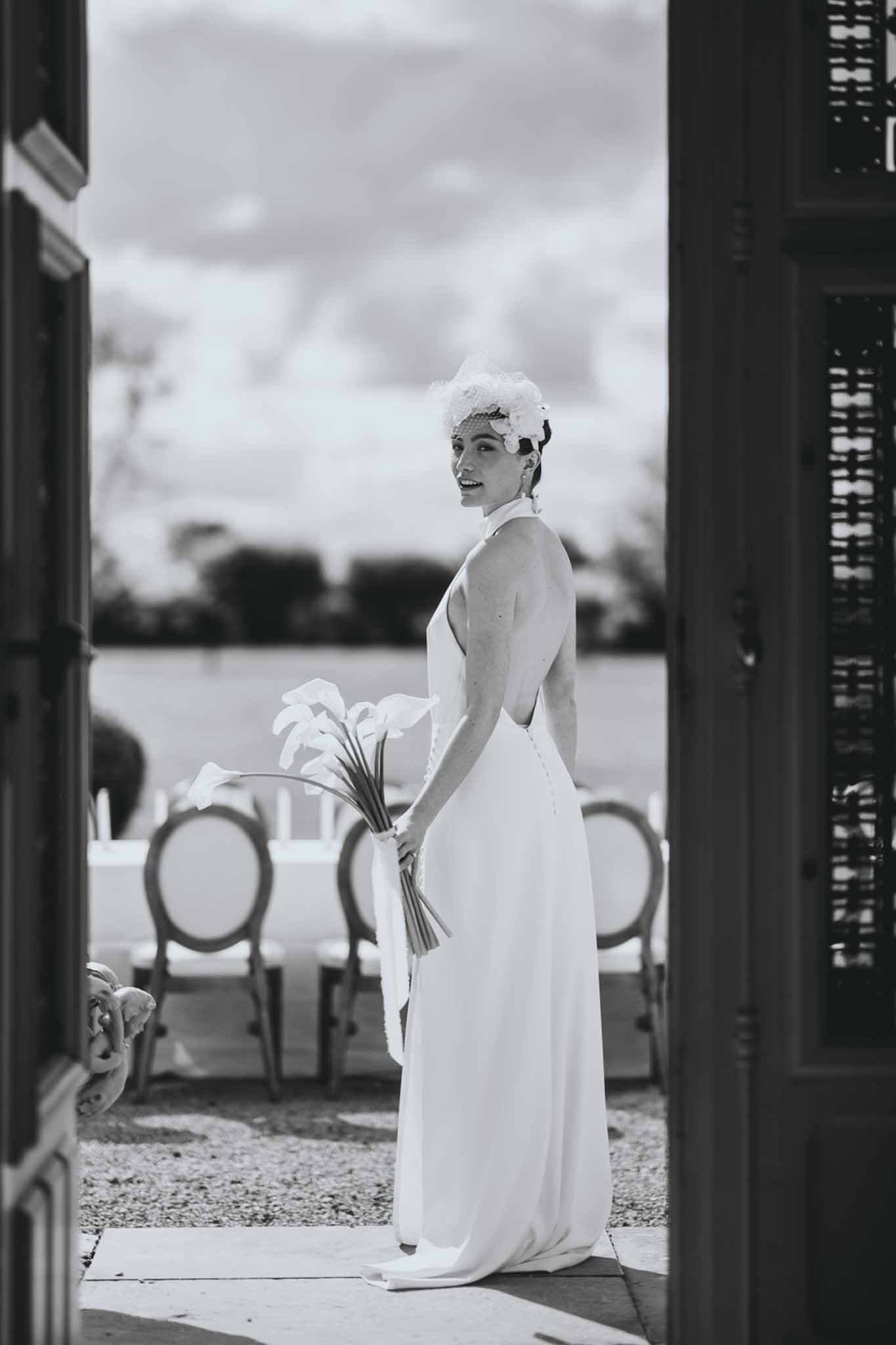Black and white portrait of bride in halter-neck gown looking over shoulder in chateau doorway