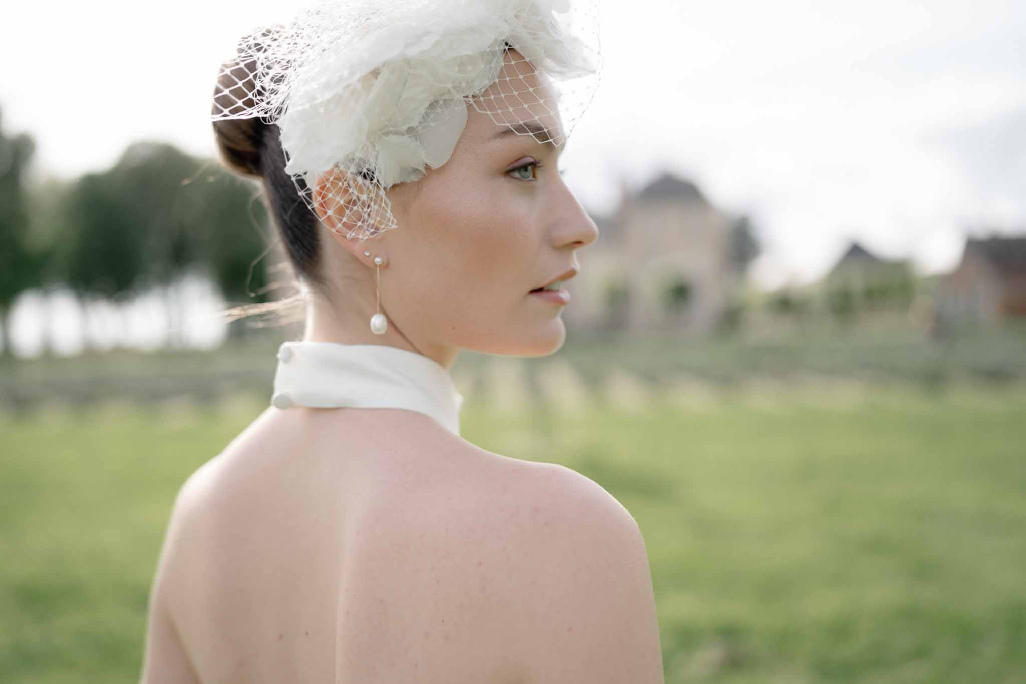 Bride from behind showing backless halter-neck gown, birdcage fascinator with tulle flowers, and pearl drop earrings