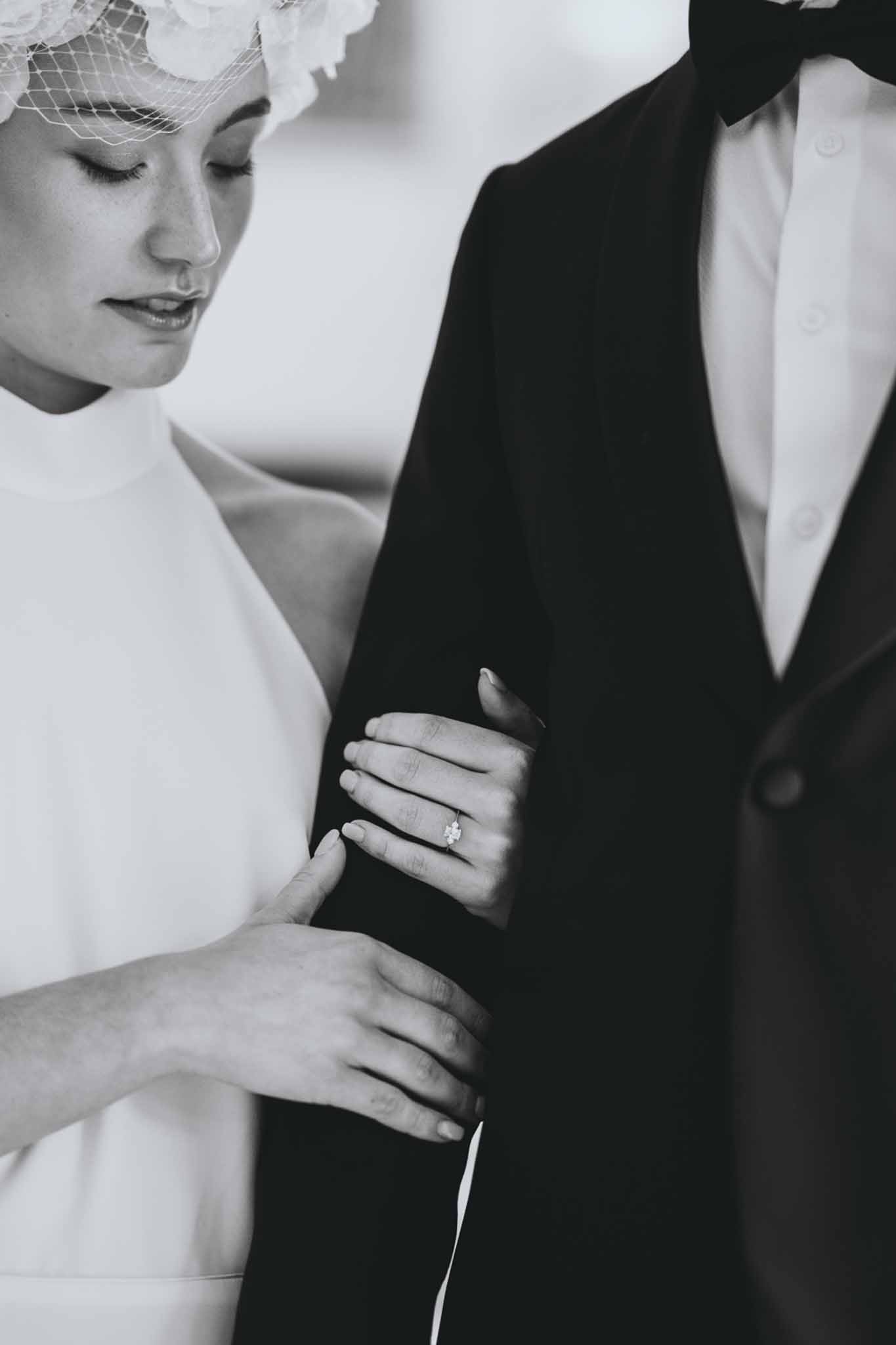 Close-up of bride with birdcage veil and solitaire ring resting hands on groom's arm in B&W