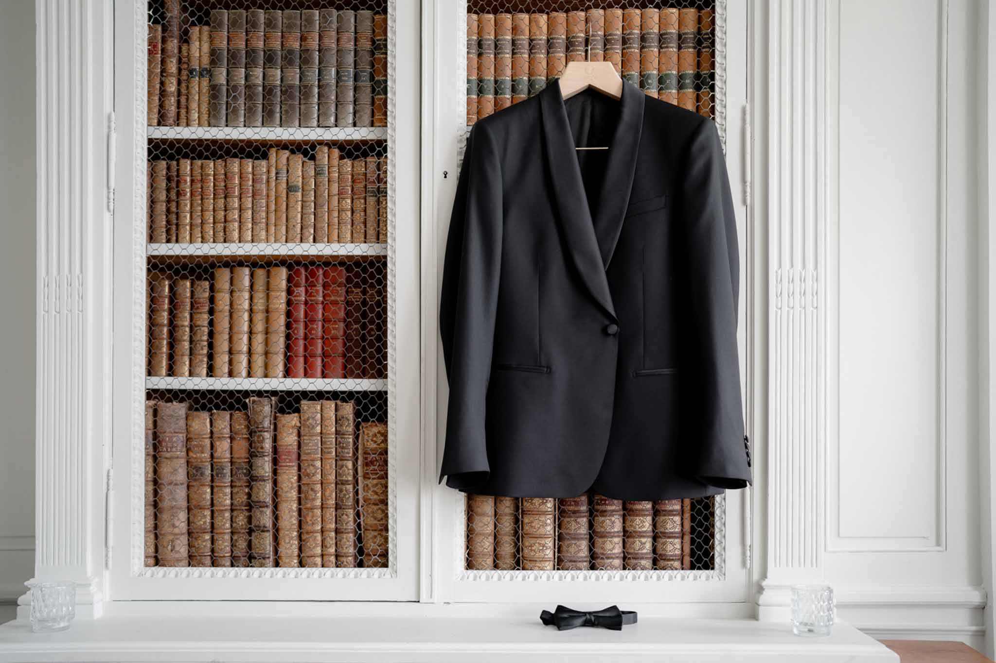 Black tuxedo jacket hung on antique bookcase with leather-bound books and silk bow tie on shelf ledge