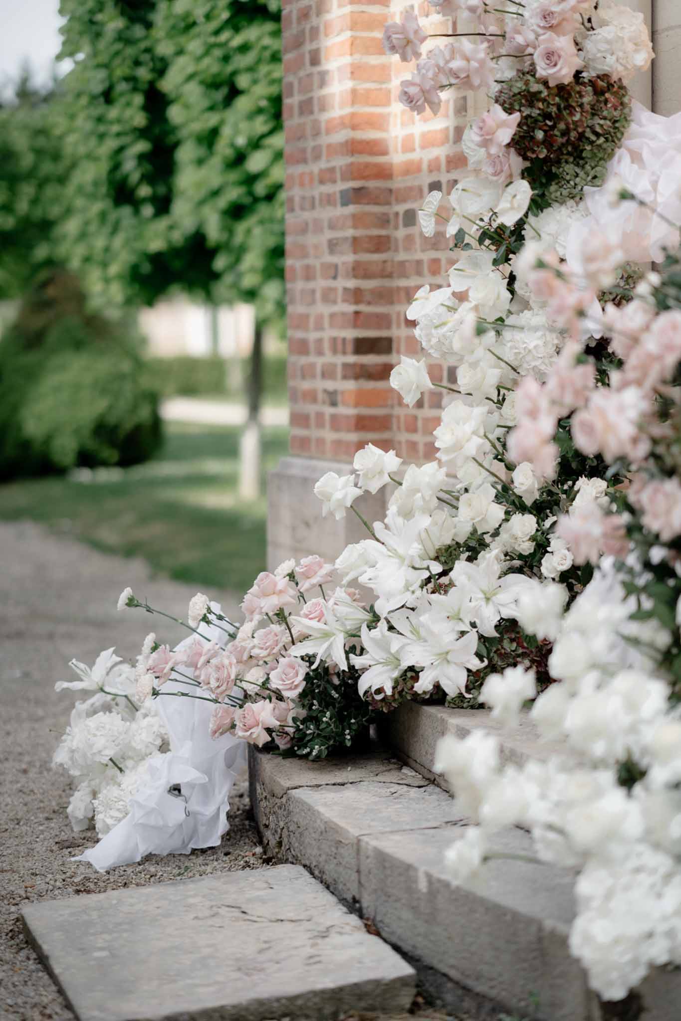 Cascading floral installation on red brick column with white lilies, blush roses, and green hydrangeas on stone steps