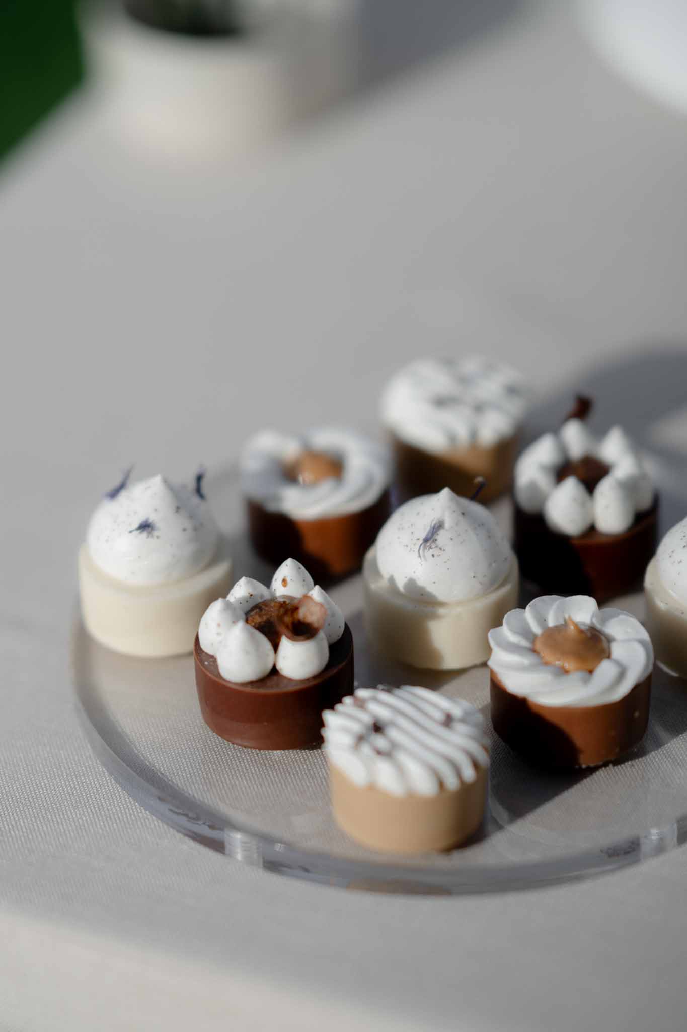 Artisan chocolate bonbons with piped cream toppings and purple speckled detail on clear acrylic stand on white linen
