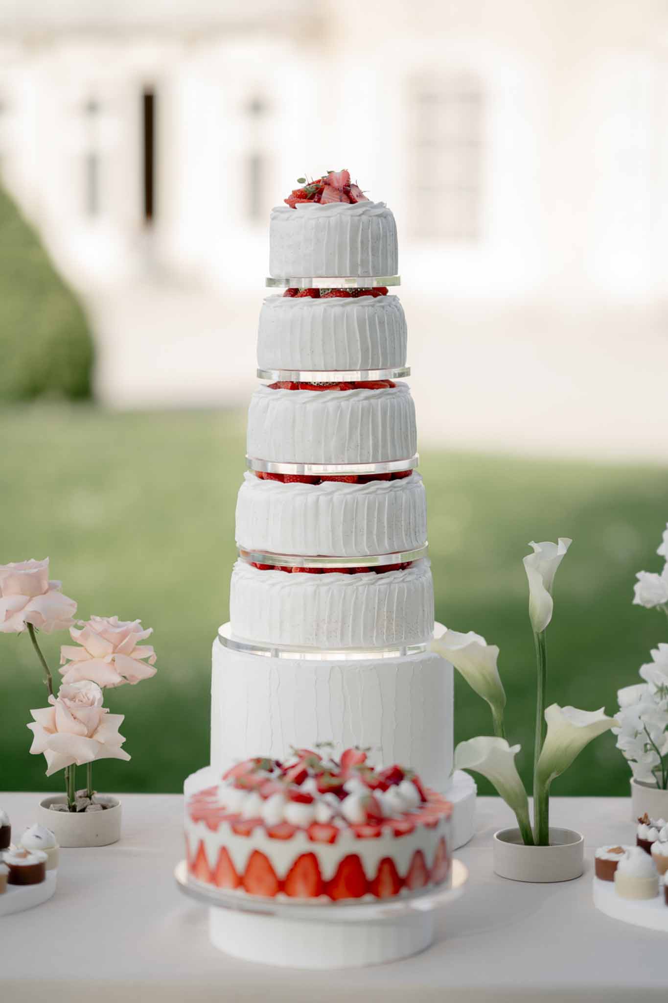 Six-tier white ruffled wedding cake with strawberry layers on acrylic spacers and fraisier cake on outdoor table