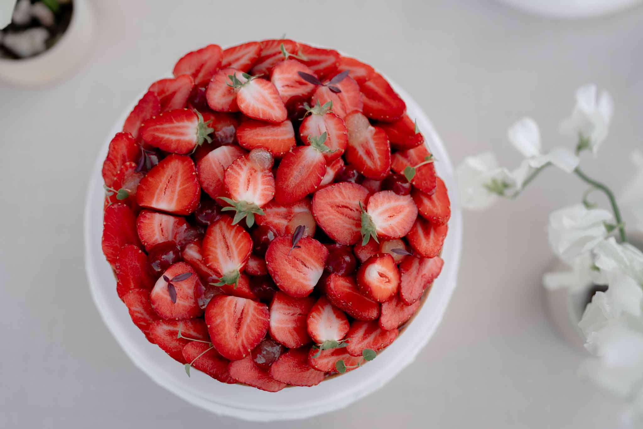 Overhead close-up of a fresh strawberry tart on a white cake stand garnished with thyme and micro-petals