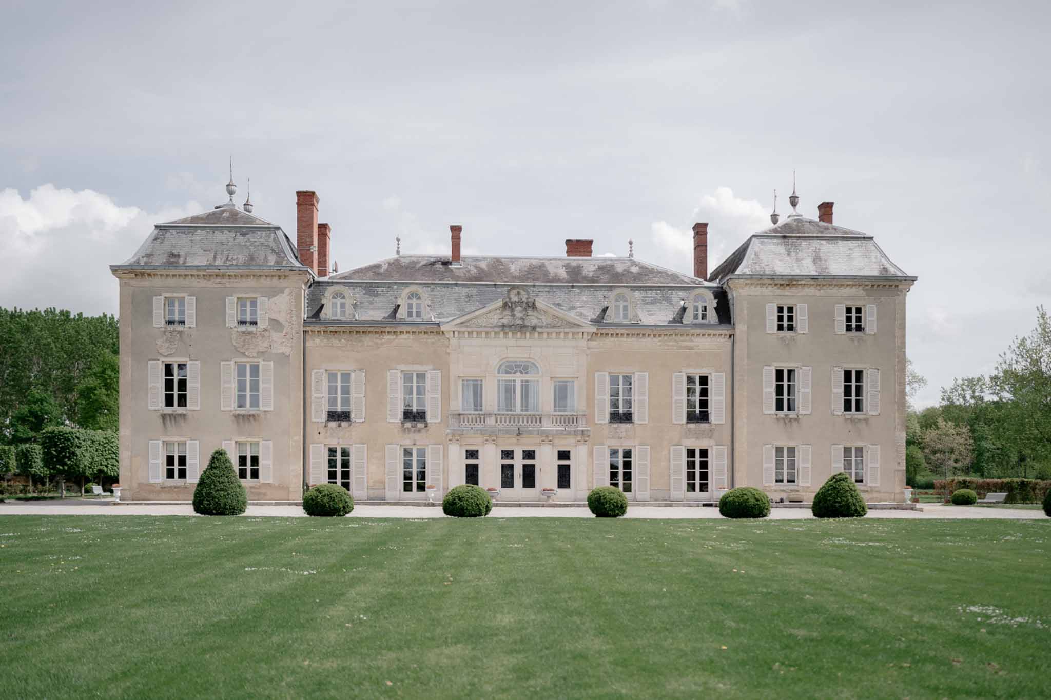 Sand-stone chateau with slate mansard roofs, carved pediment, and box topiary spheres on gravel terrace