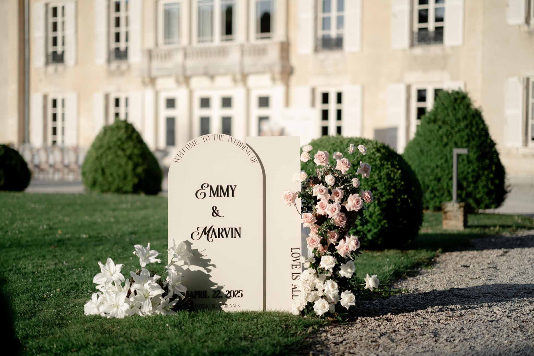 Cream arch welcome signs with blush rose cascade and white lilies before chateau with topiary garden