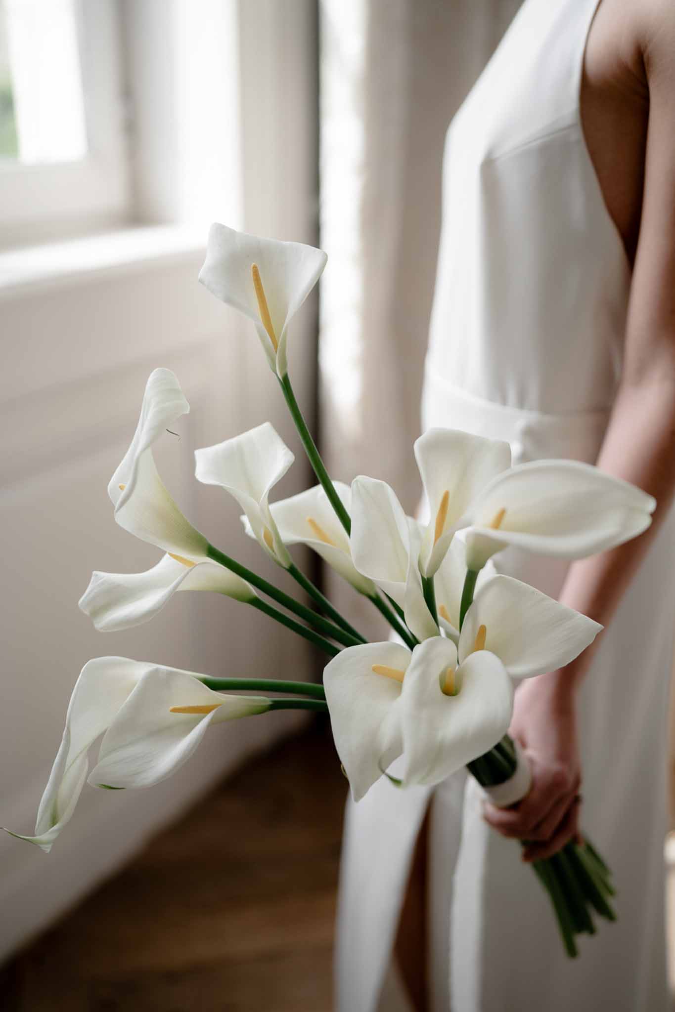 Bride holding minimalist bouquet of white calla lilies with ribbon wrap in getting-ready room