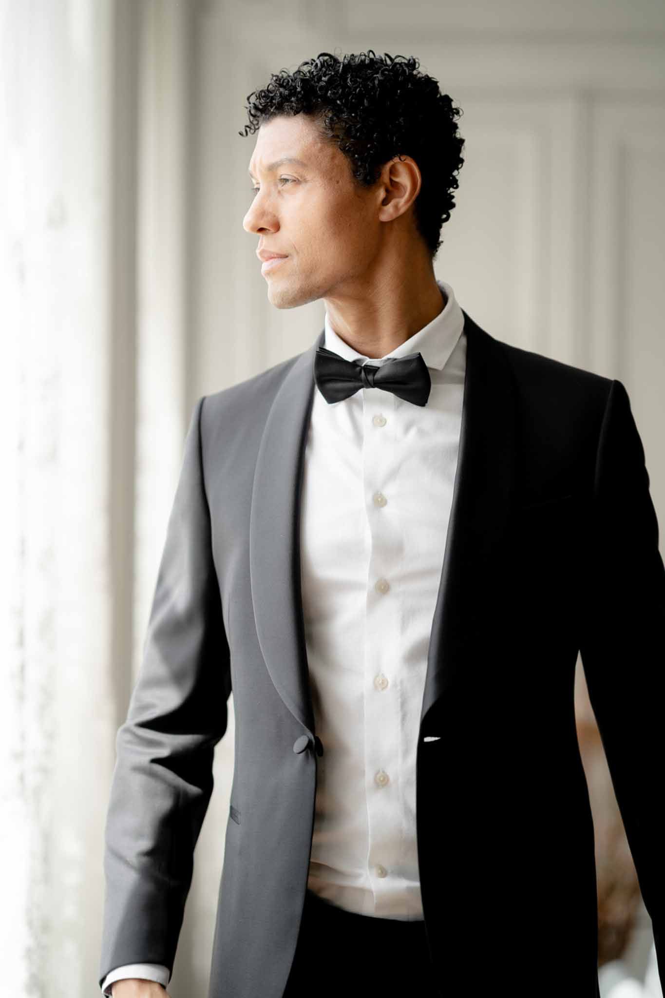 Groom in dark grey tuxedo with satin lapels and black bow tie gazing to side in chateau interior