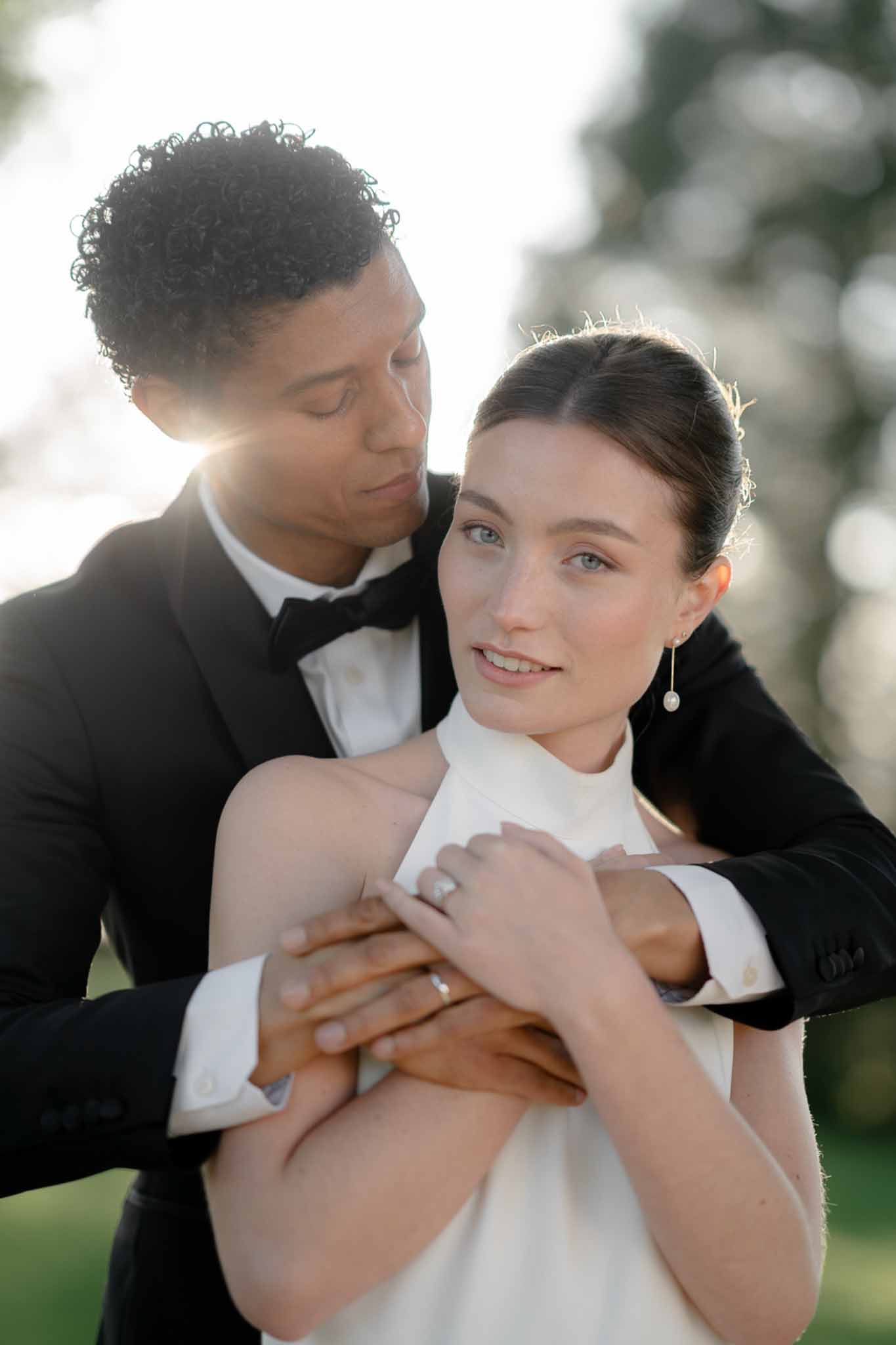 Groom in black tuxedo embracing bride in white halter-neck gown during golden hour backlit portrait