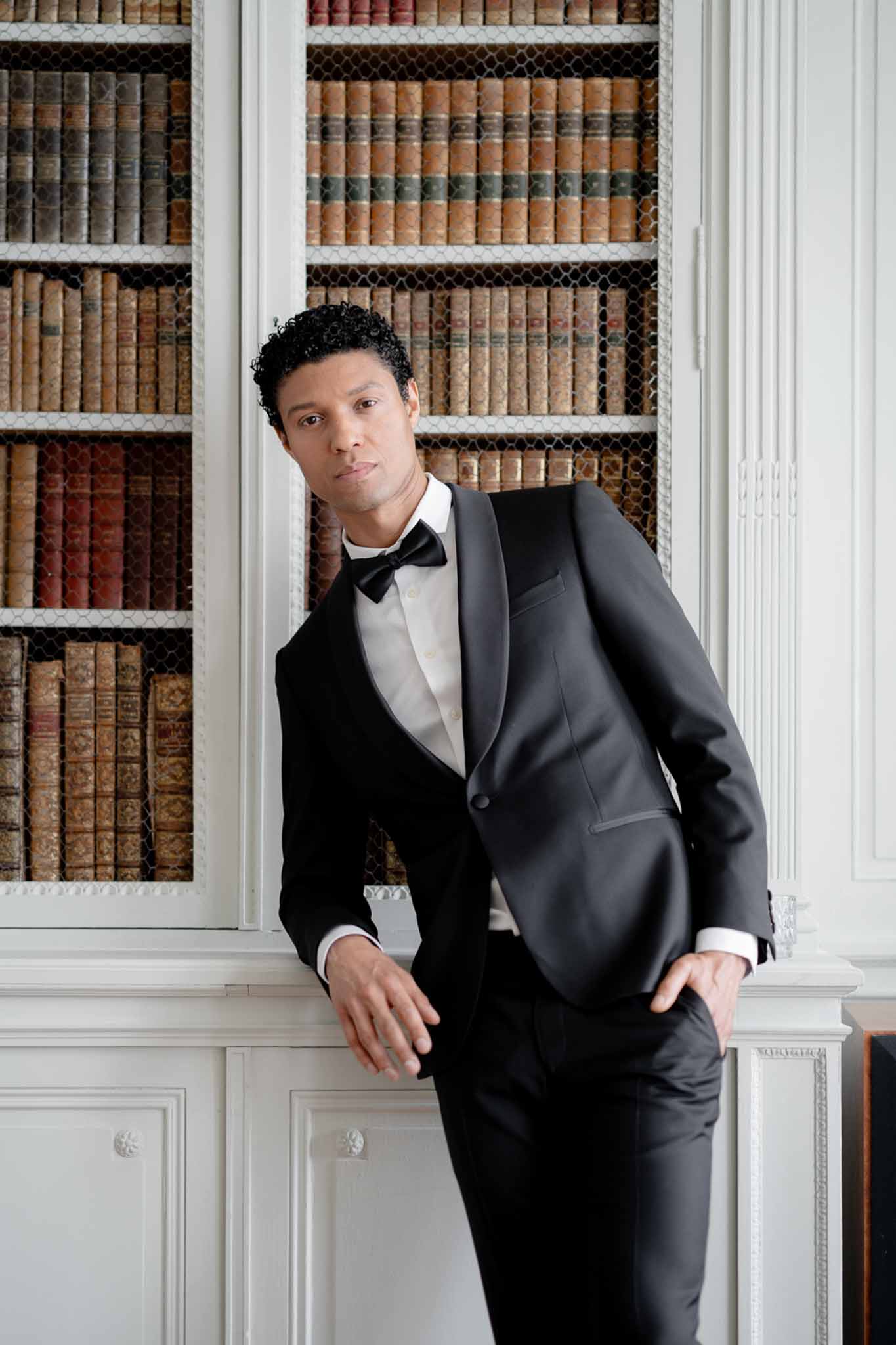 Groom in black satin-lapel tuxedo leaning against bookcase of antique leather-bound volumes