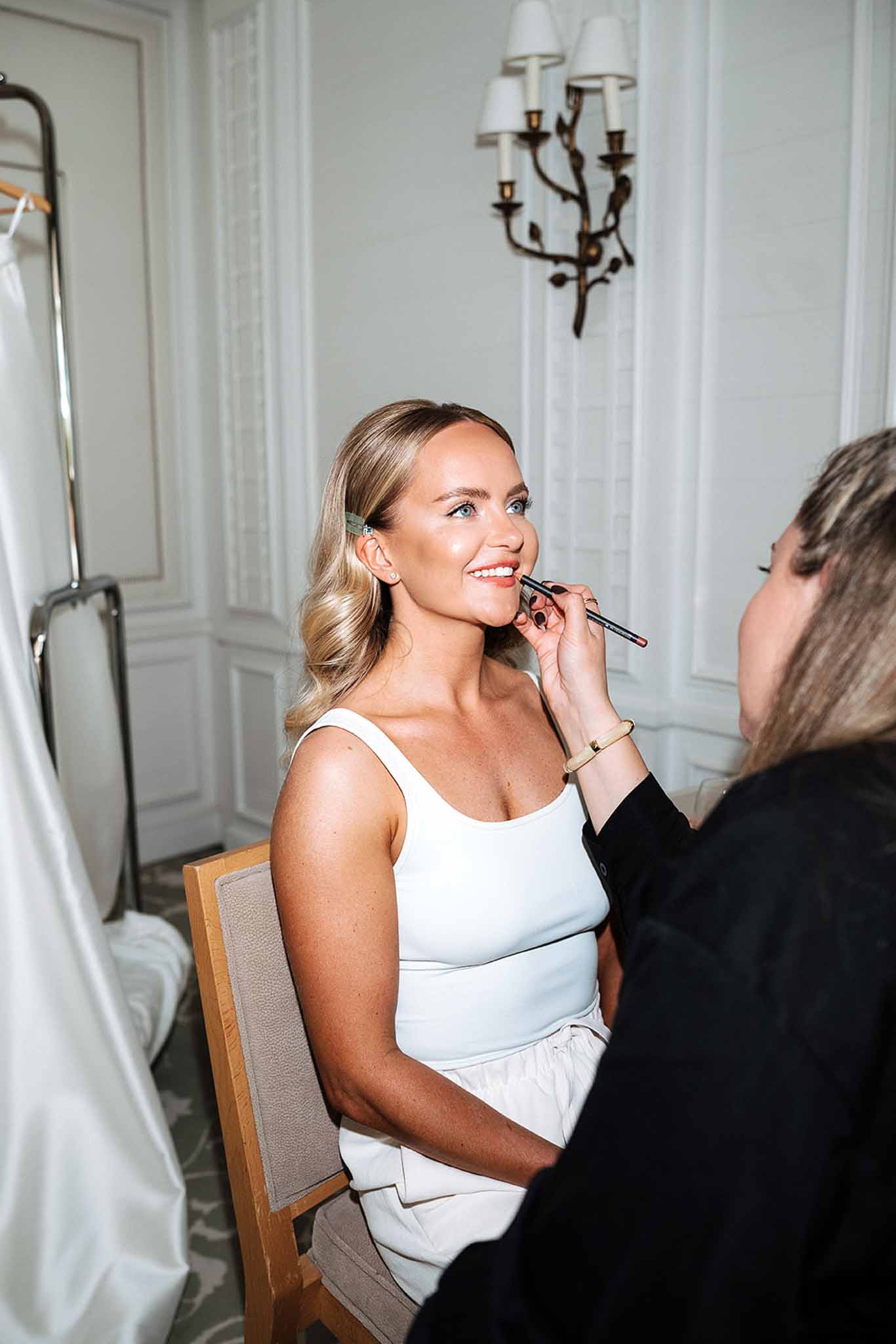 Makeup artist applying lipstick to smiling bride seated in chateau bridal suite with white paneled walls