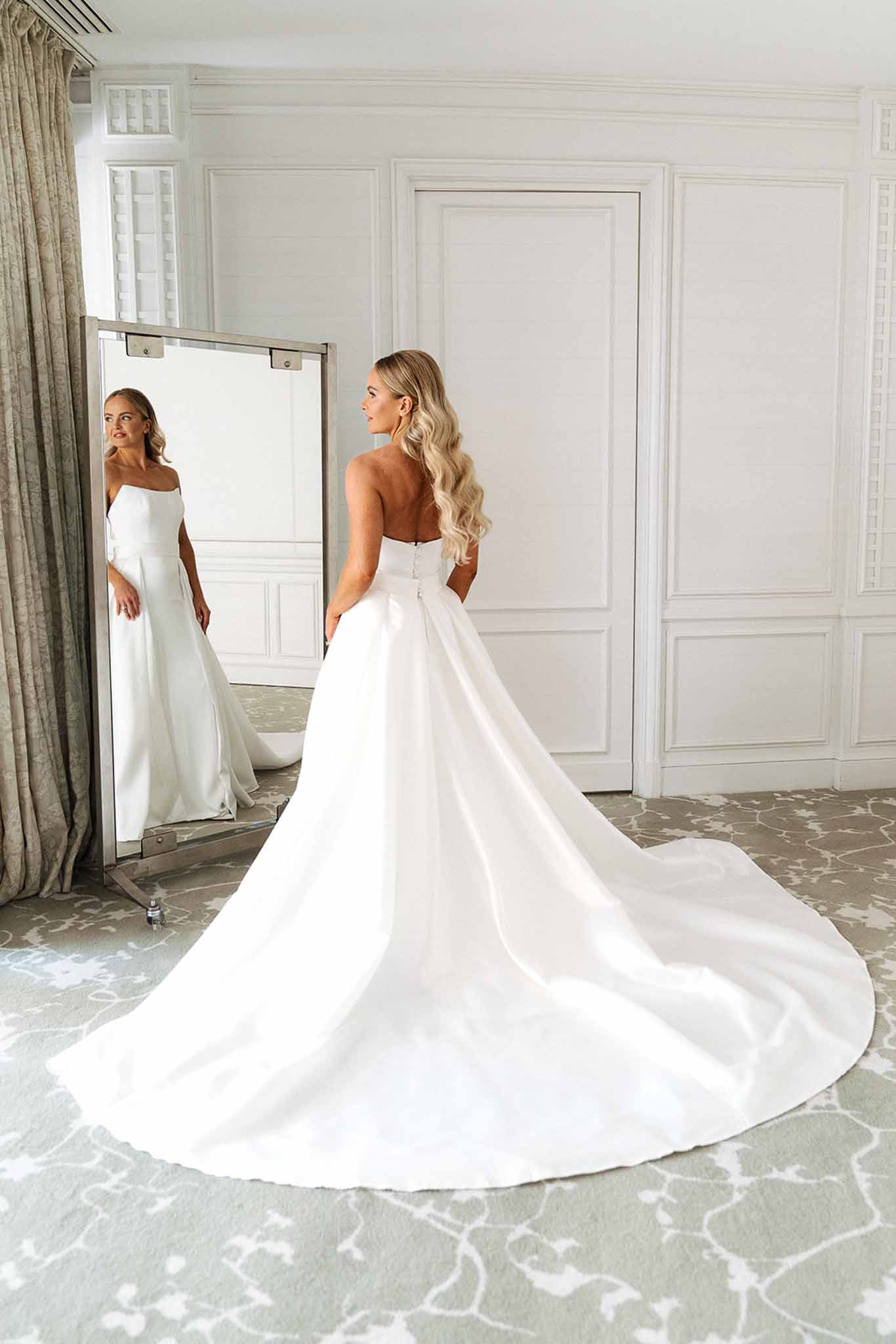 Bride seen from behind in strapless white satin ball gown with cathedral train, looking into freestanding mirror