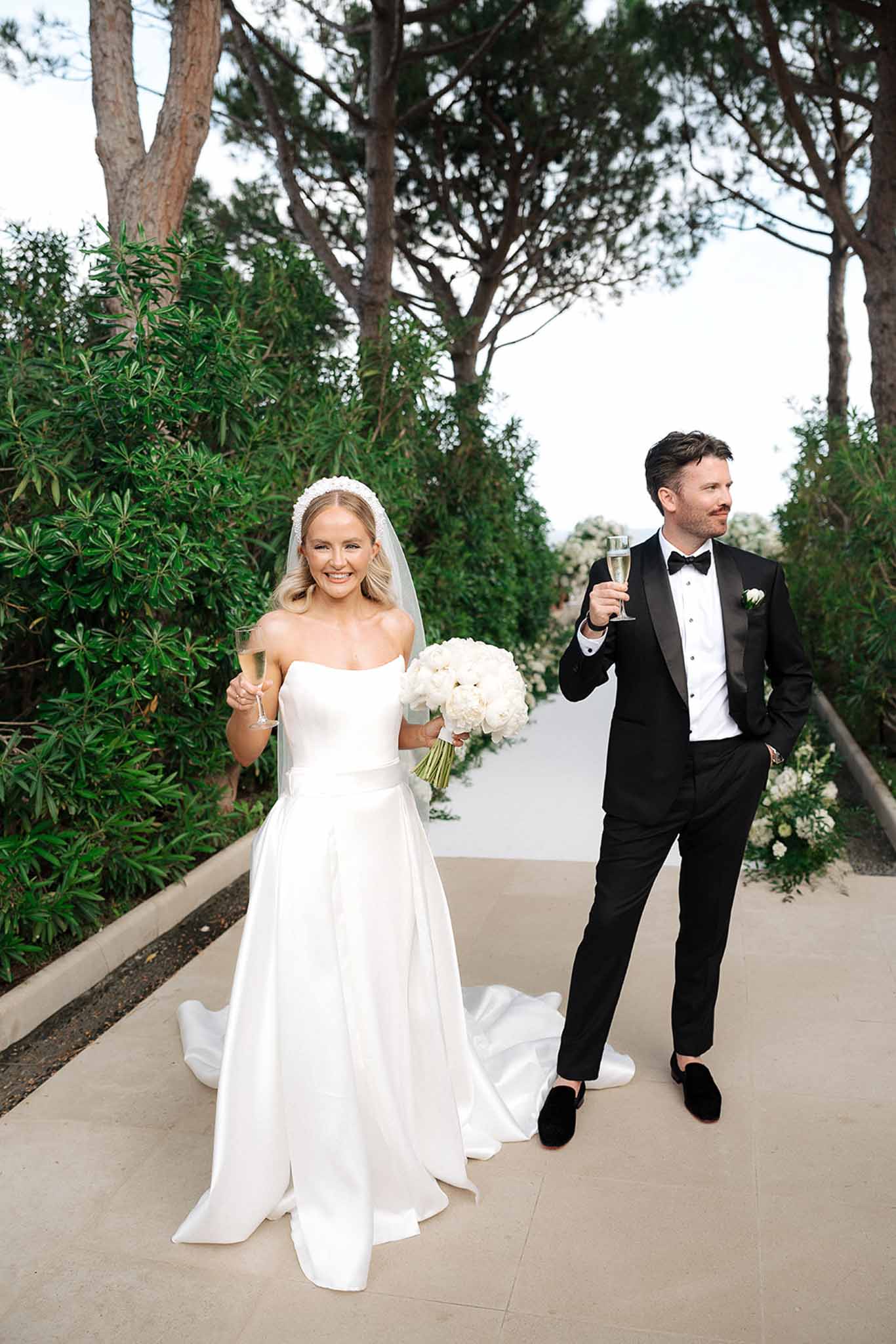 Bride with pearl headband and white peony bouquet raising champagne flute beside groom in tuxedo on terrace