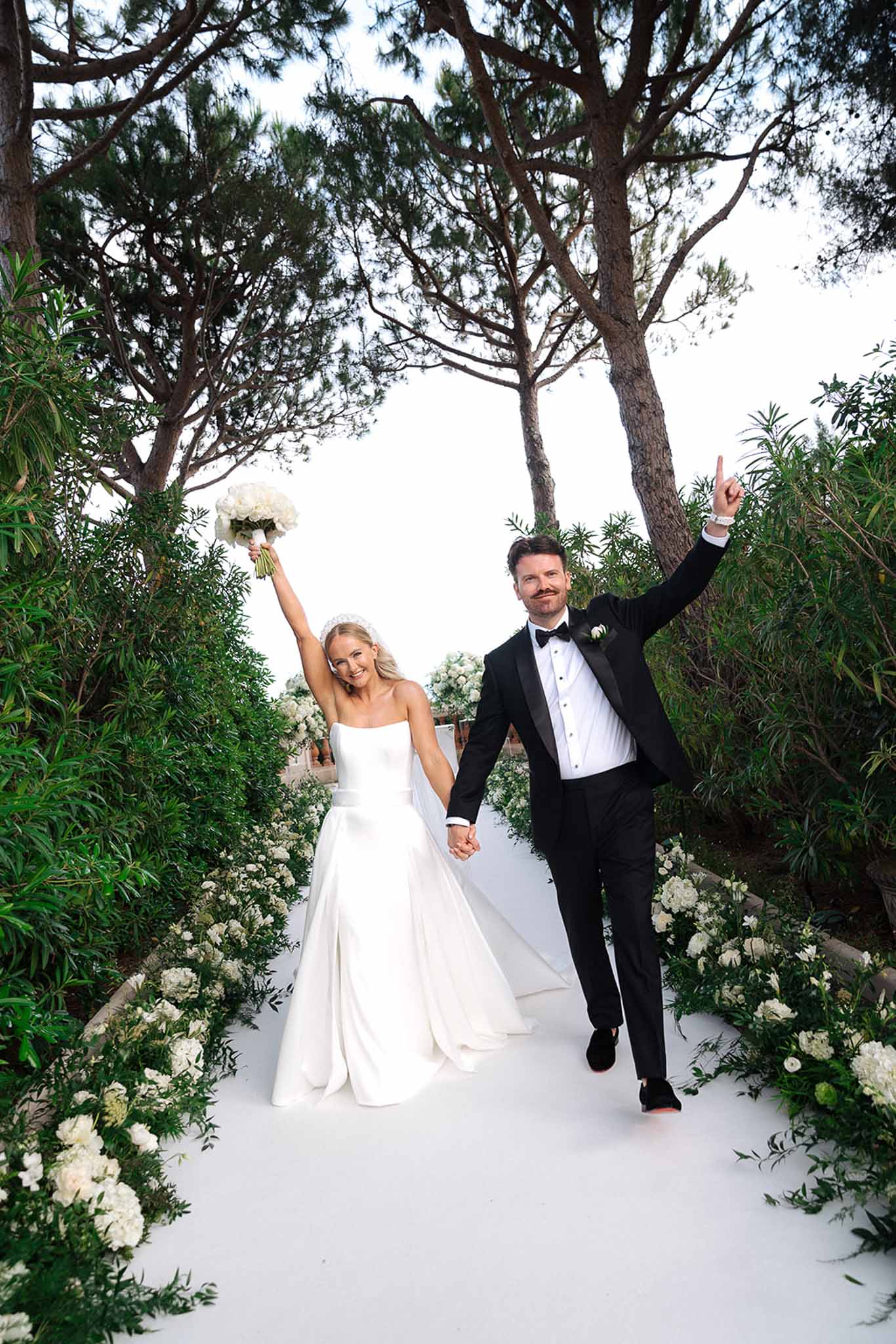 Couple raising arms in celebration walking down white hydrangea-lined aisle after ceremony