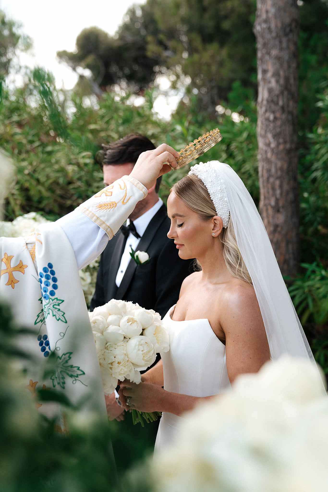 Priest placing gold stefana crown on bride with white peony bouquet during outdoor Orthodox ceremony