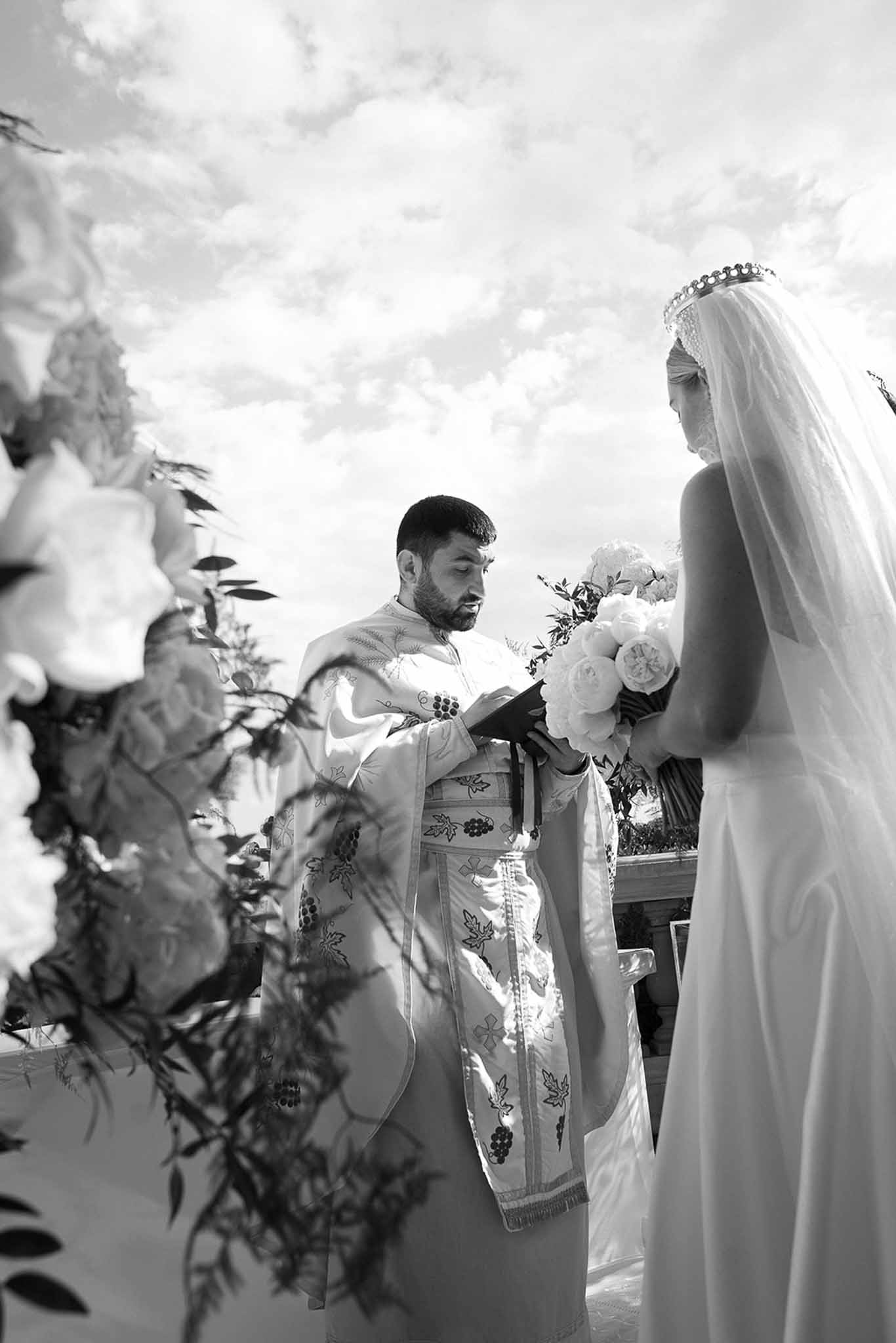 Black and white image of priest reading to bride in strapless gown and veil during outdoor religious ceremony