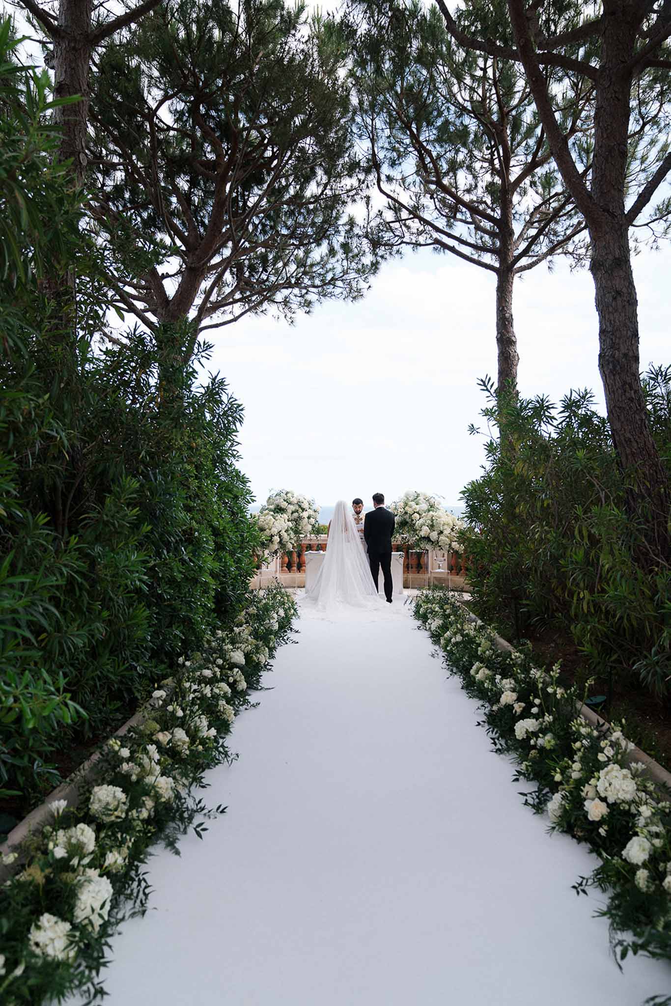 Outdoor ceremony view down white aisle lined with white peonies and hydrangeas toward couple at terrace altar