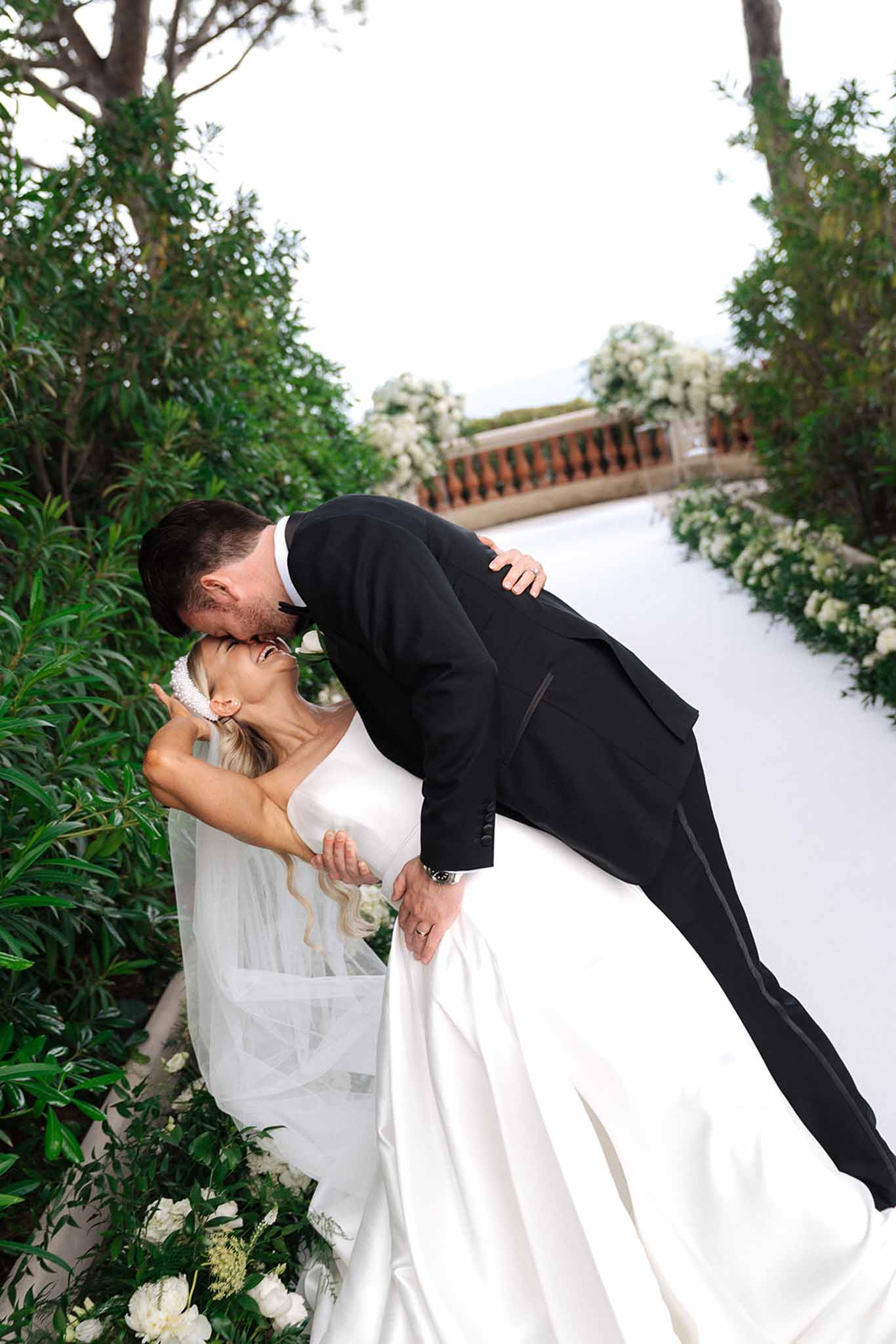 Bride and groom share dip kiss on ceremony aisle lined with white hydrangea and rose arrangements at stone terrace