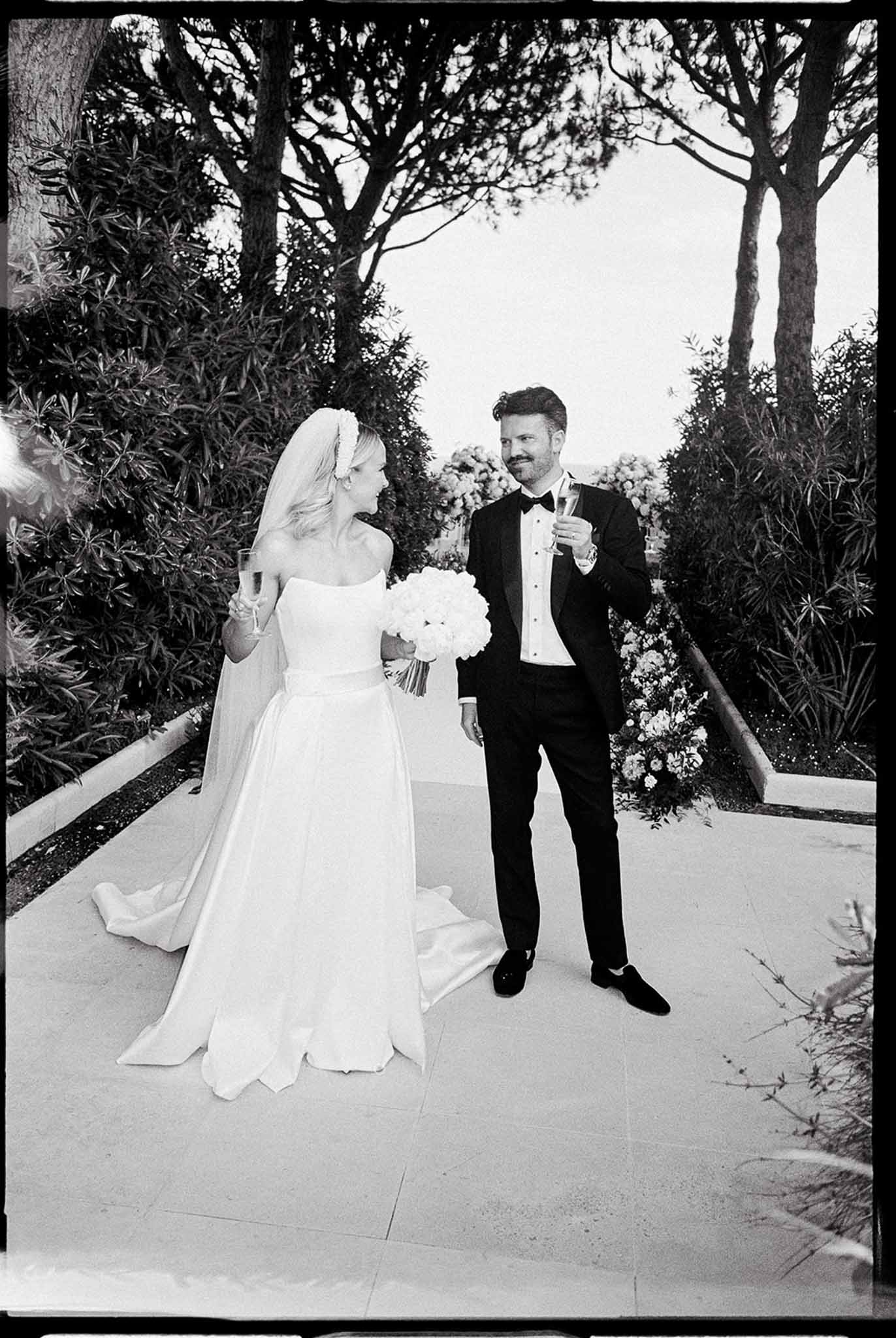 Black-and-white film photo of bride in ballgown and groom in tuxedo holding champagne on stone terrace