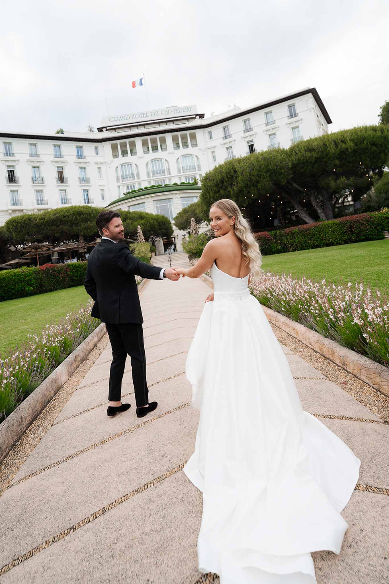 Bride in strapless ballgown leading groom along lavender-bordered path toward Belle Epoque hotel facade