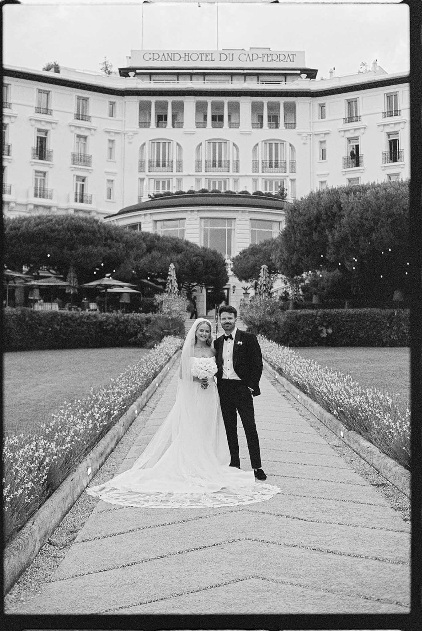 Black and white bride and groom on path before Grand Hotel du Cap Ferrat facade
