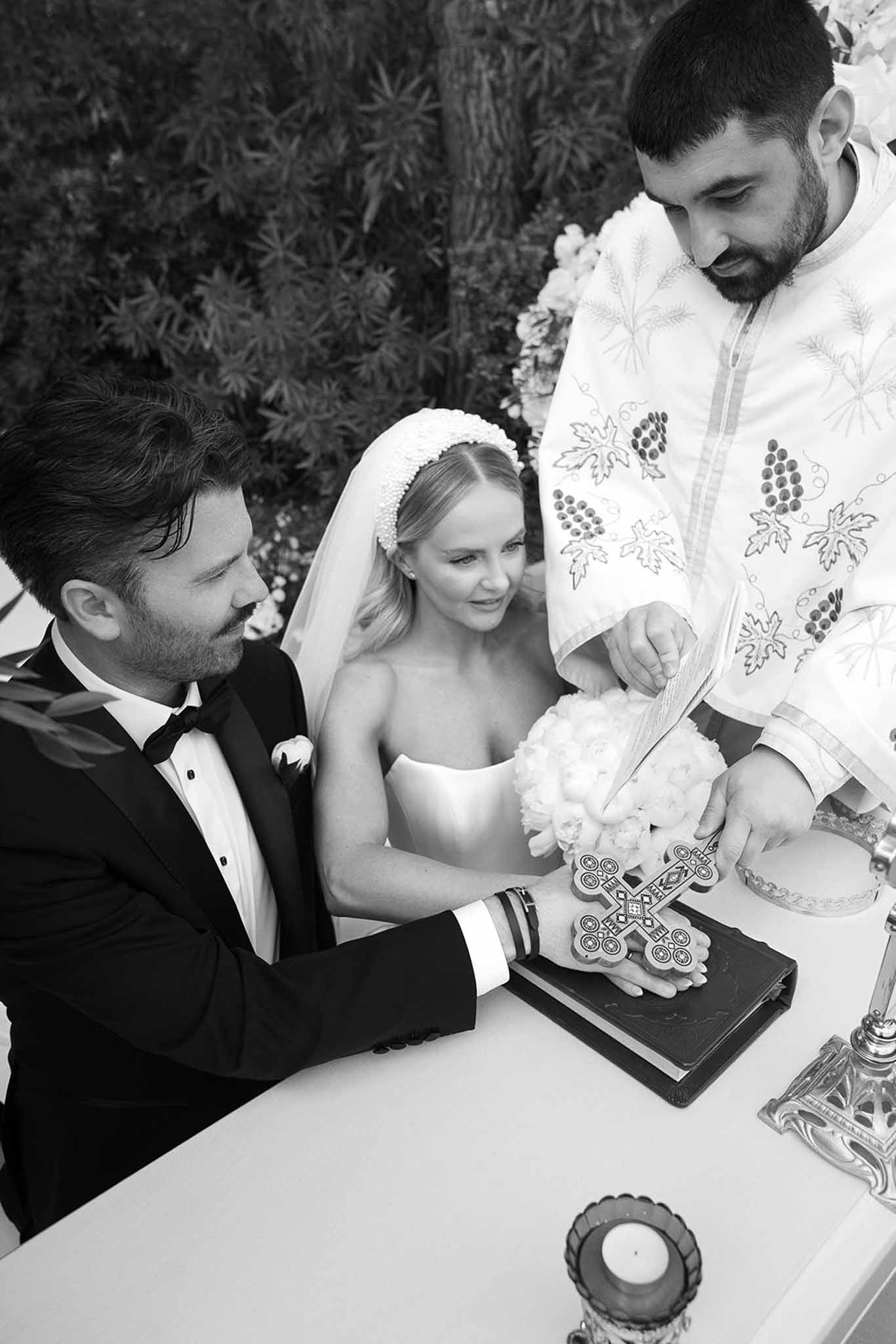 Black and white close-up Orthodox ceremony priest placing cross on Bible grooms hand bride with pearl headband and bouquet