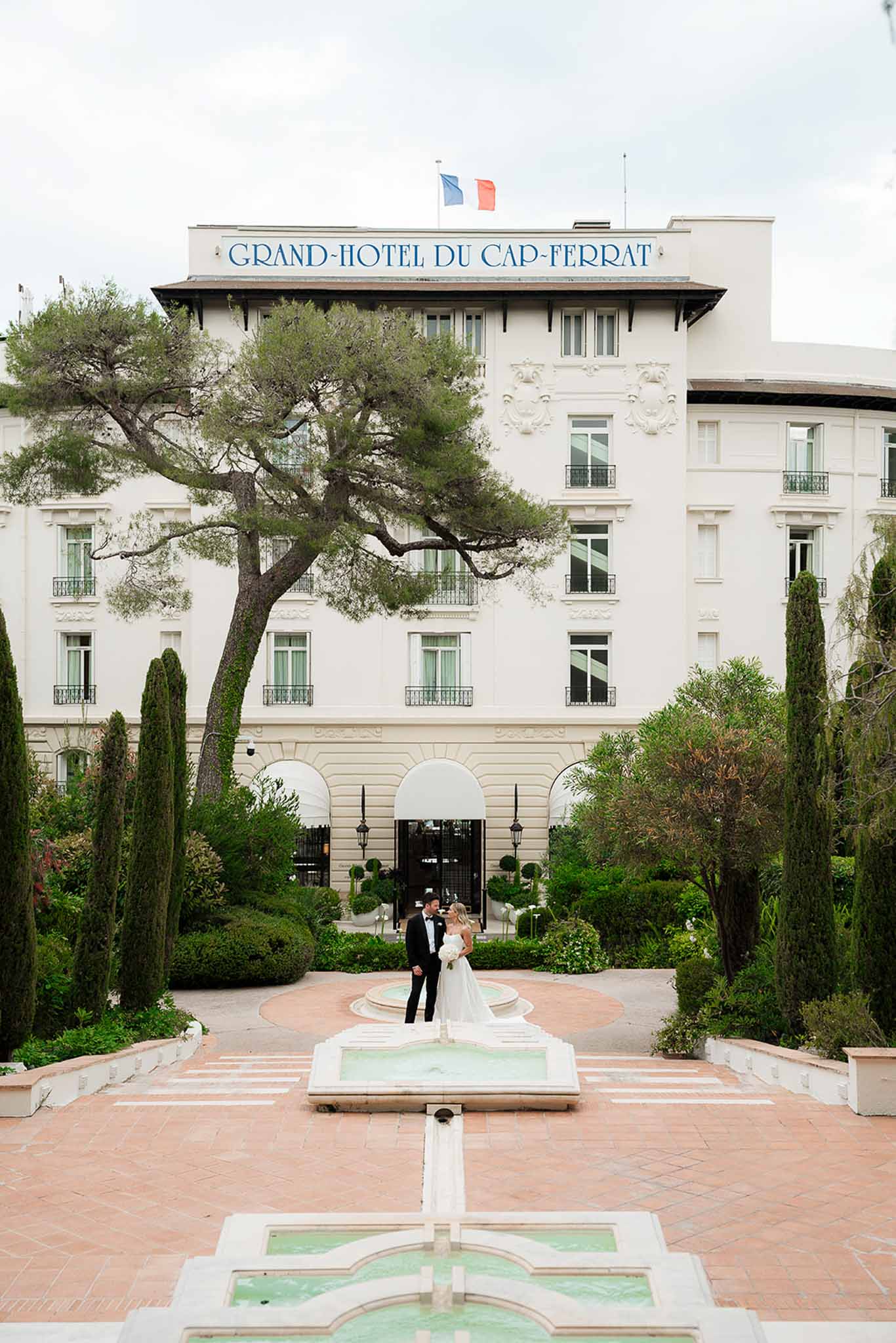 Bride and groom standing by fountain at Grand-Hotel du Cap-Ferrat with formal gardens and hotel facade