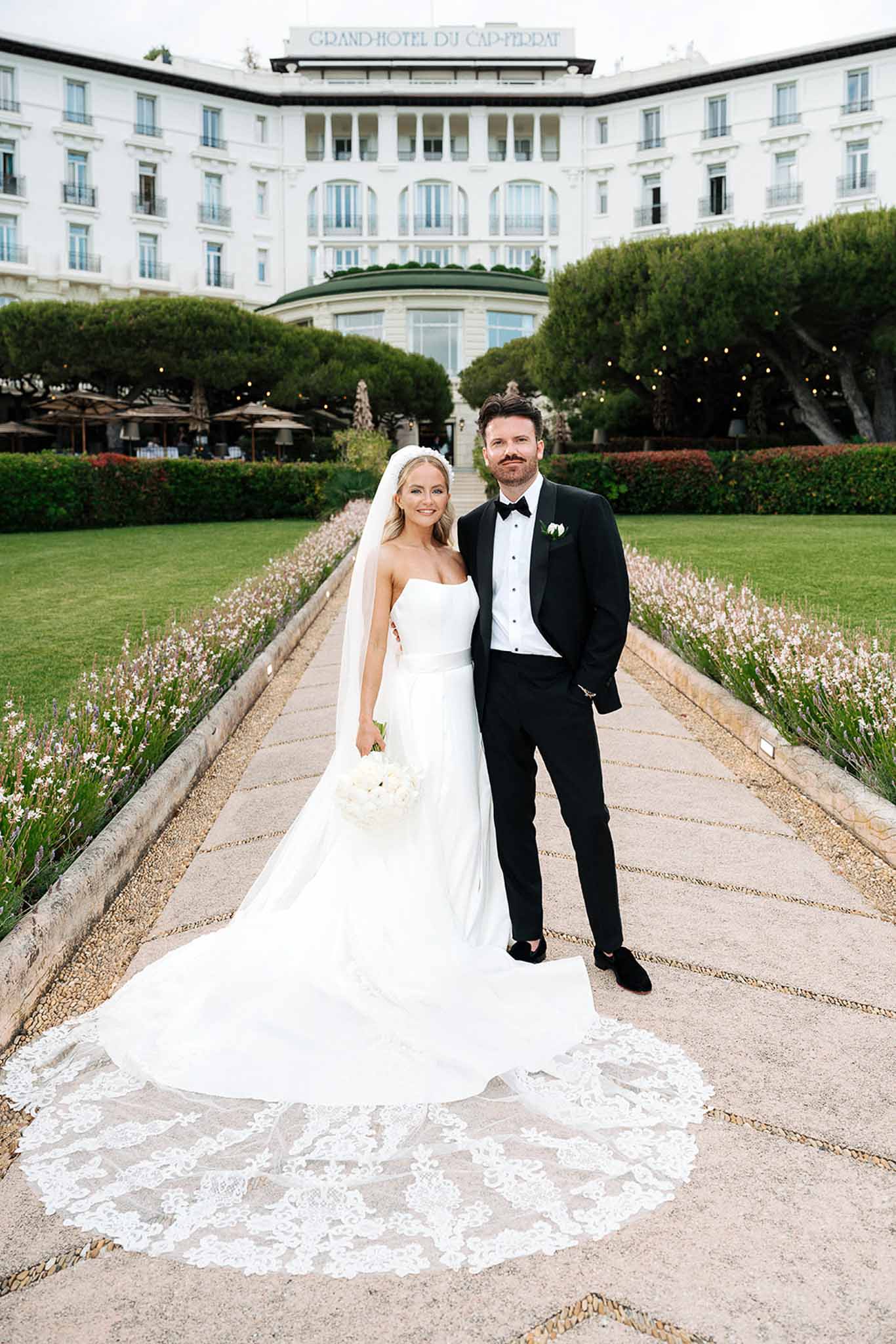 Couple on gravel path at Belle Epoque hotel, bride in lace-bordered cathedral veil with white peony bouquet