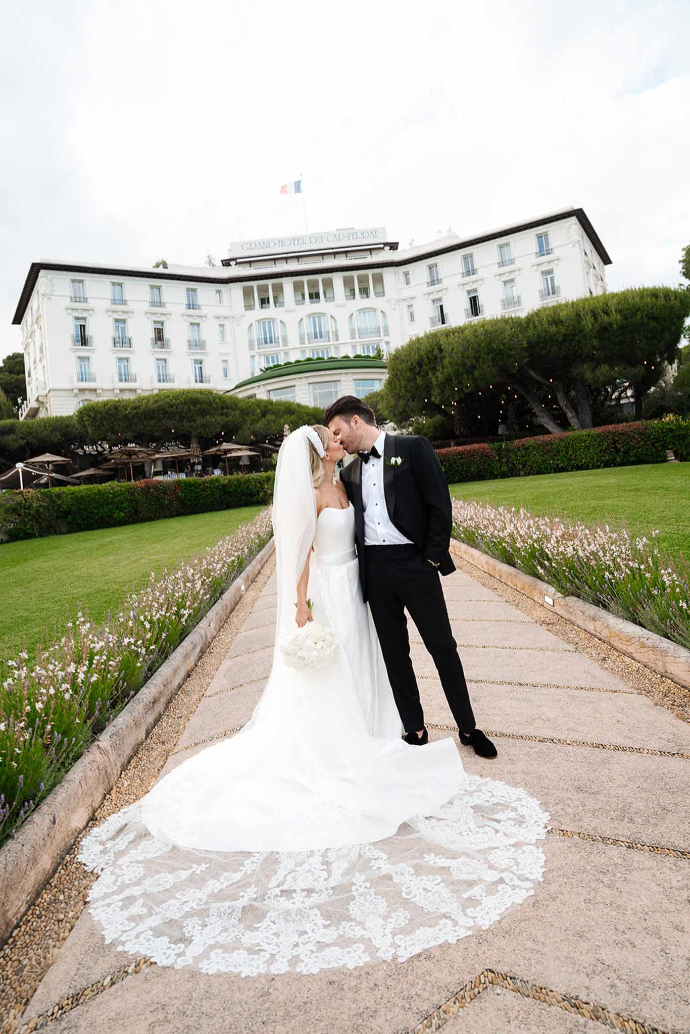 Bride and groom kissing on gravel path at Grand-Hotel du Cap-Ferrat with cathedral lace veil spread across walkway