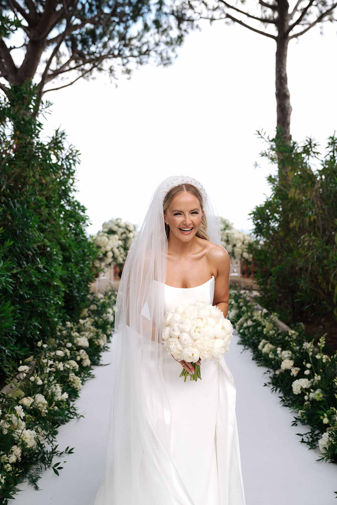Bride laughing on white aisle in strapless satin gown and pearl headband with white ranunculus bouquet and floral border