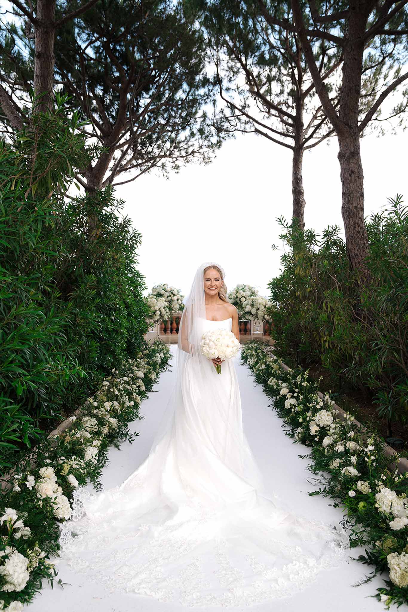 Bride in strapless white ball gown and cathedral veil holding peony bouquet on a flower-lined outdoor aisle