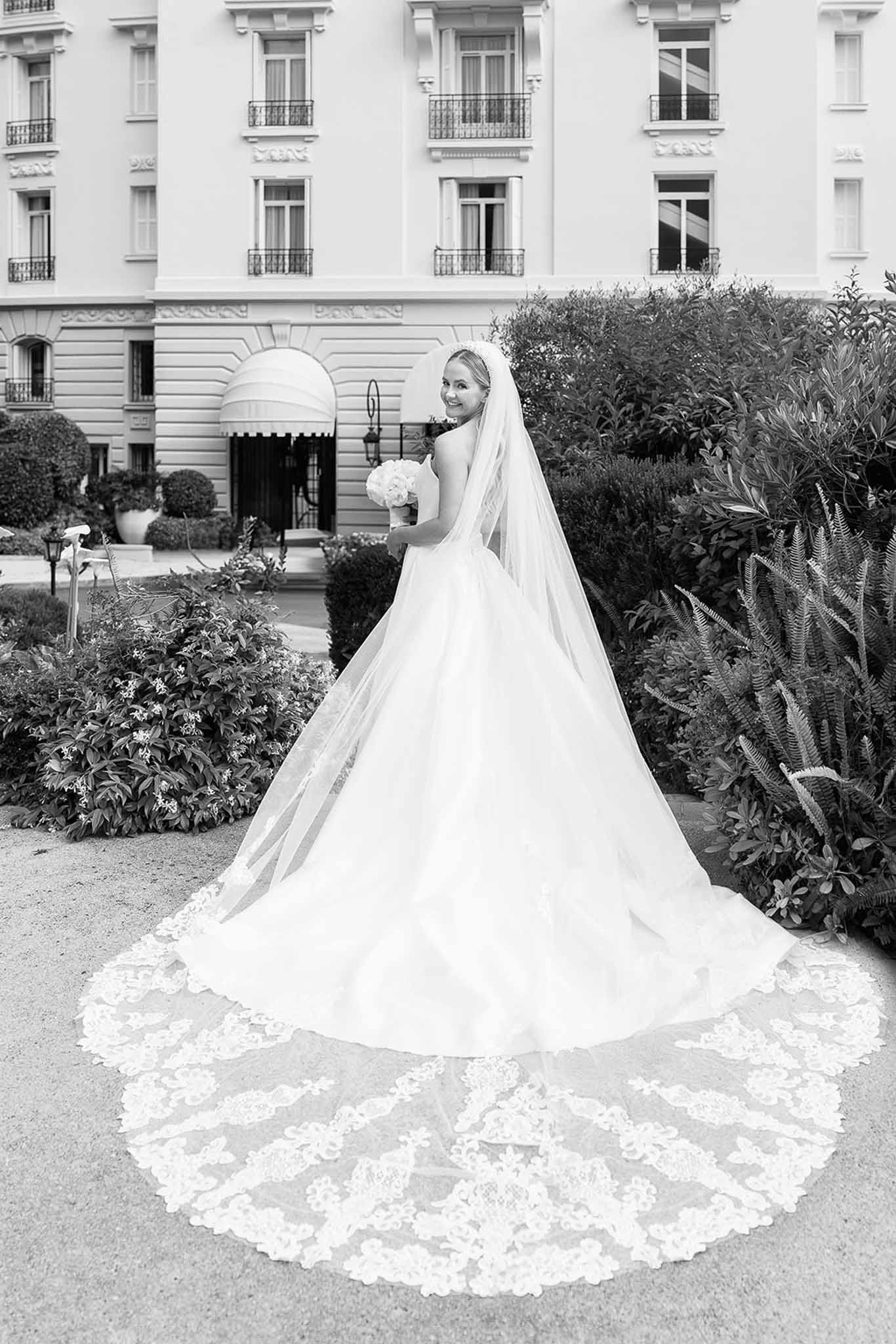 Black-and-white bridal portrait from behind showing cathedral-length lace veil fanning across gravel garden path