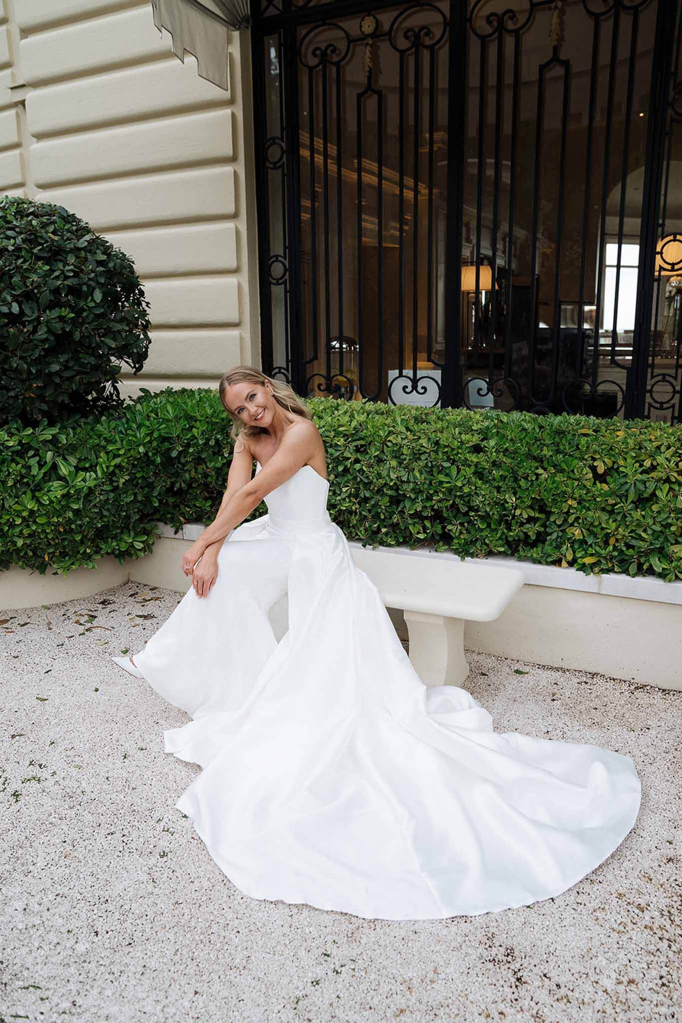 Bride in strapless white ballgown seated on stone bench with cathedral train spread across gravel courtyard