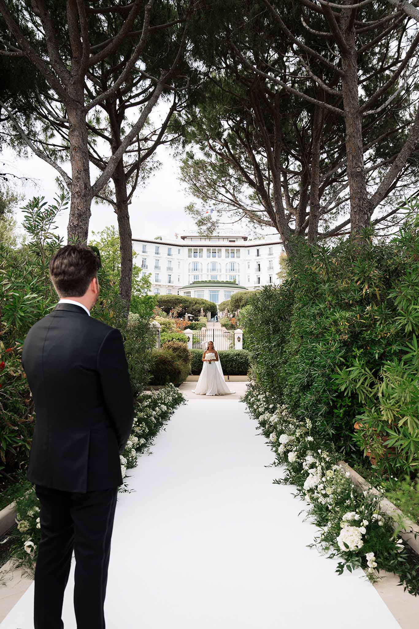Groom watching bride approach along white aisle runner lined with florals at Belle Epoque hotel