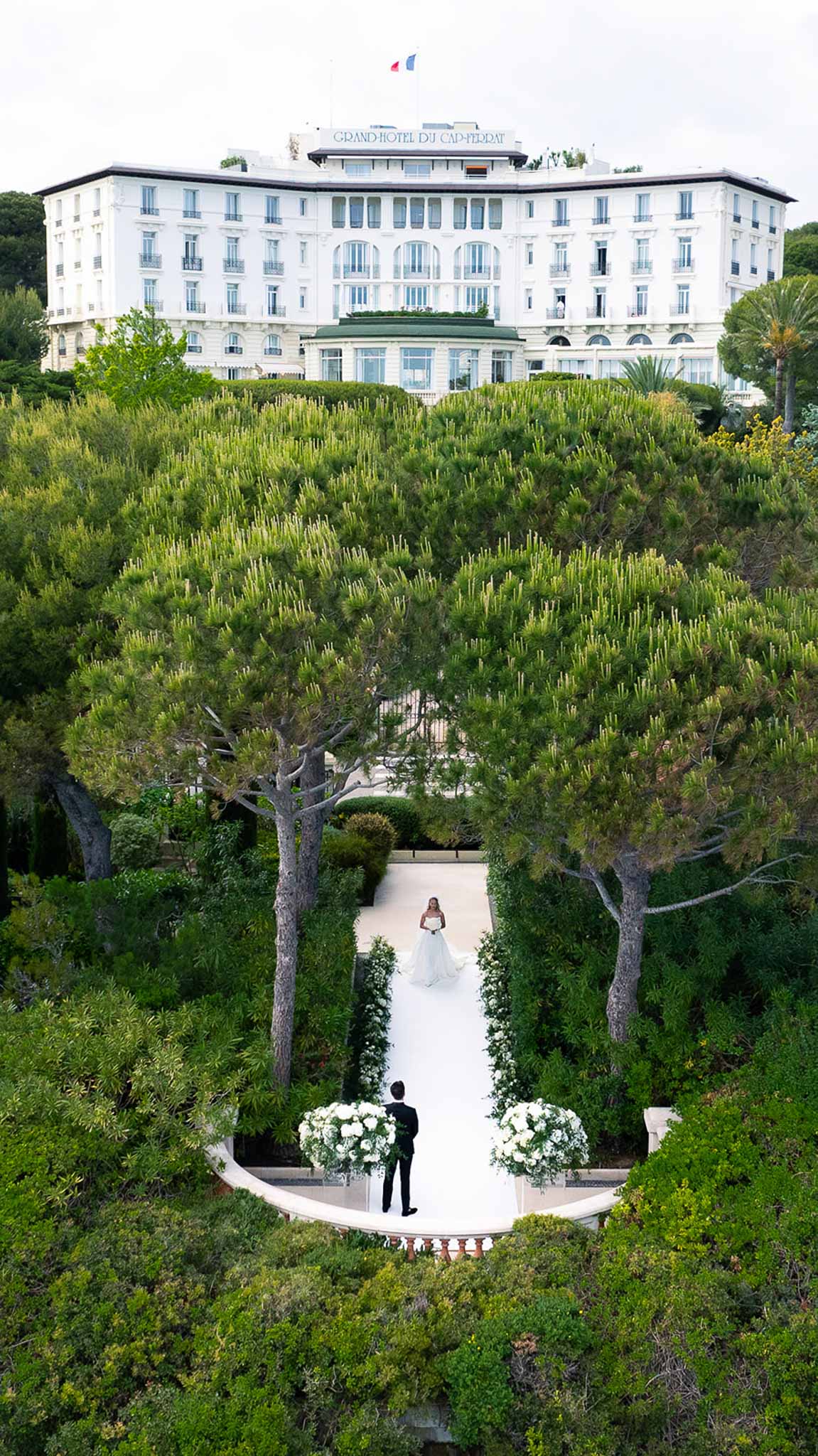Aerial view of bride walking white aisle toward groom at Grand-Hotel du Cap-Ferrat with hedgerow border