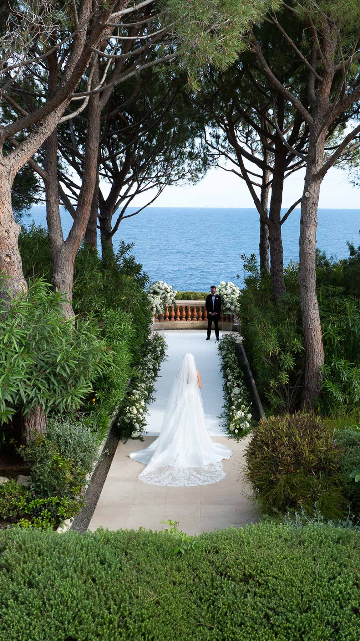 Bride walking down aisle toward groom on stone terrace overlooking the sea with white floral arrangements