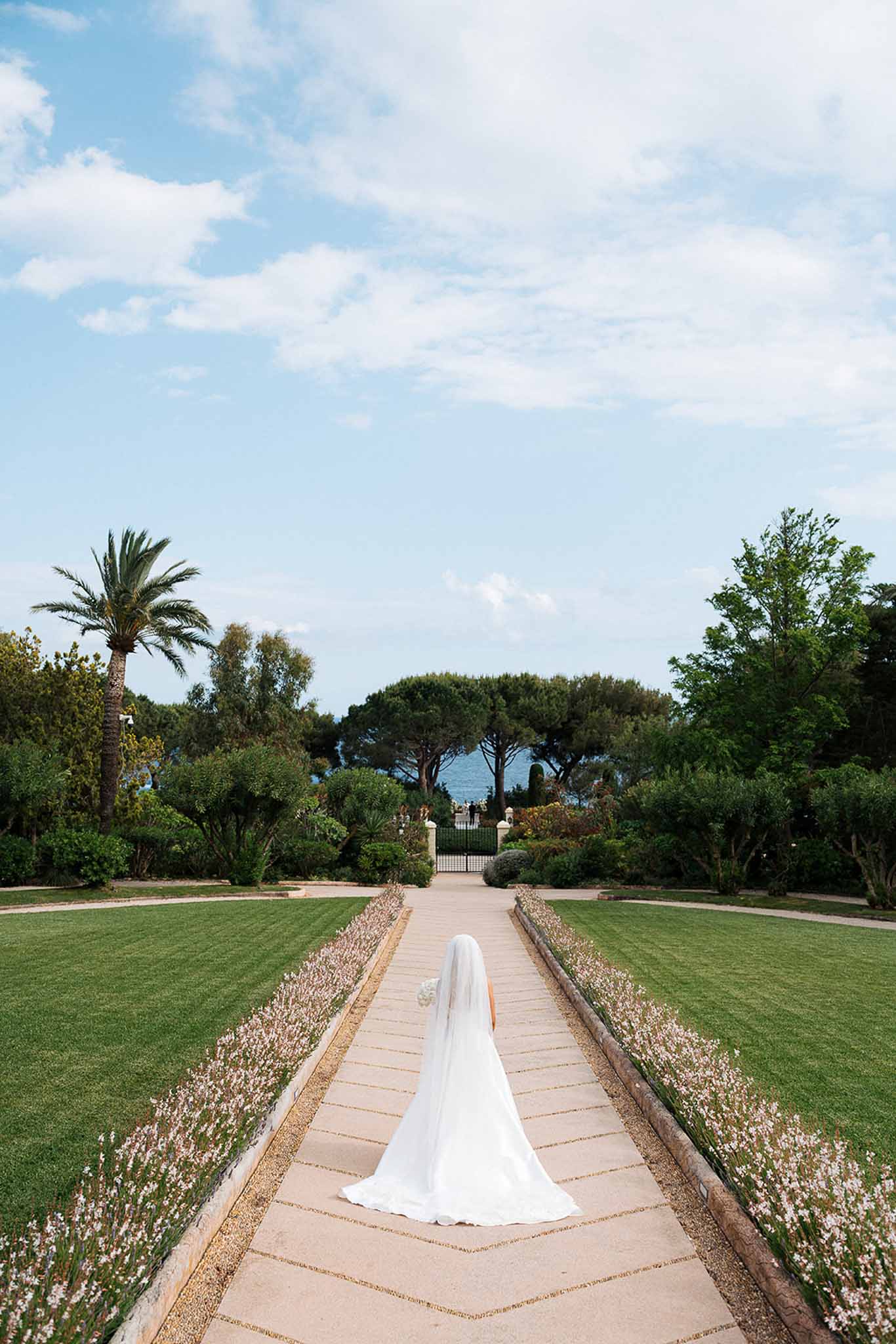 Bride walks away along lavender-bordered path with cathedral veil toward iron gates and sea view