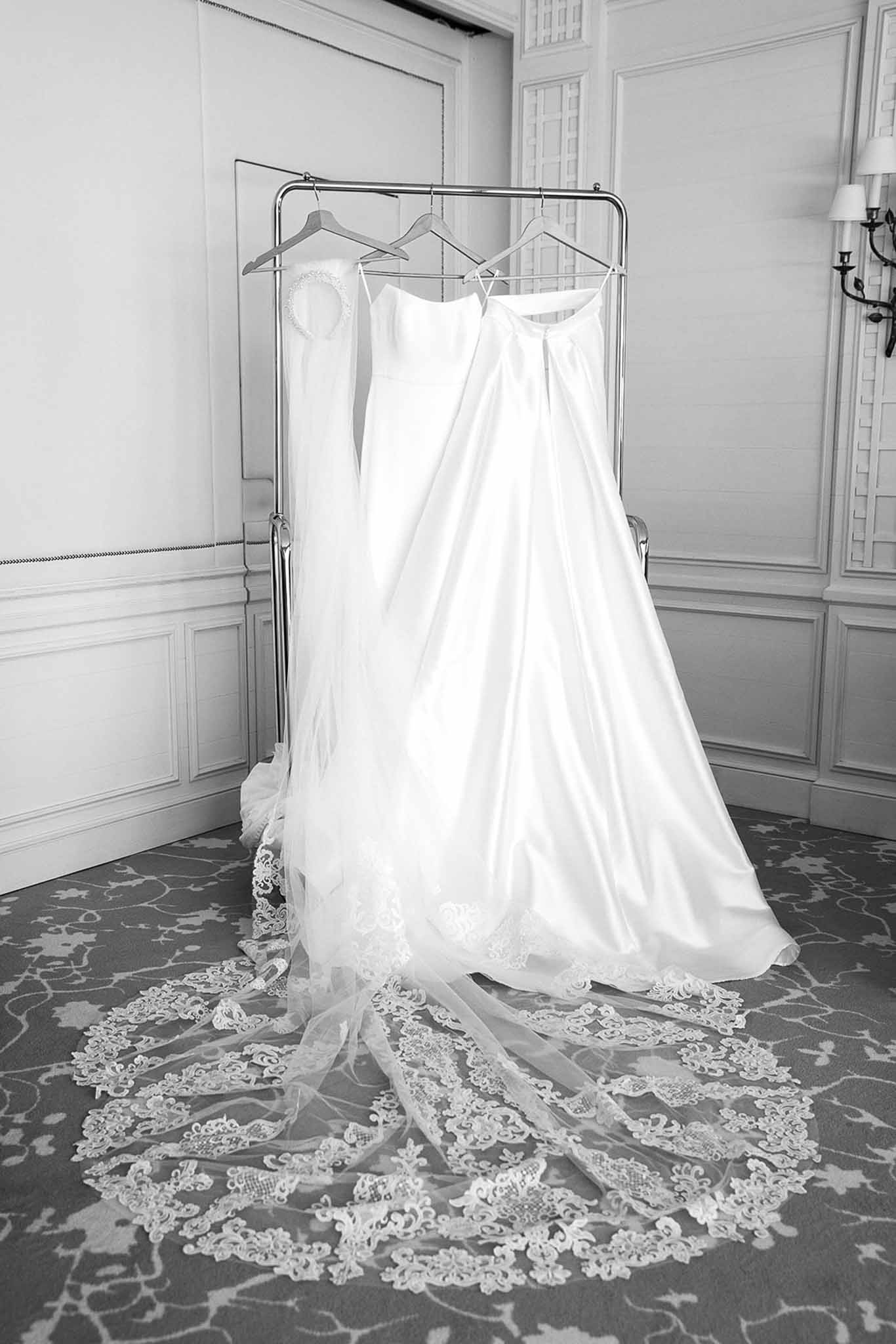 Two white wedding gowns hanging on chrome rack with cathedral-length lace veil spread on patterned carpet in black and white