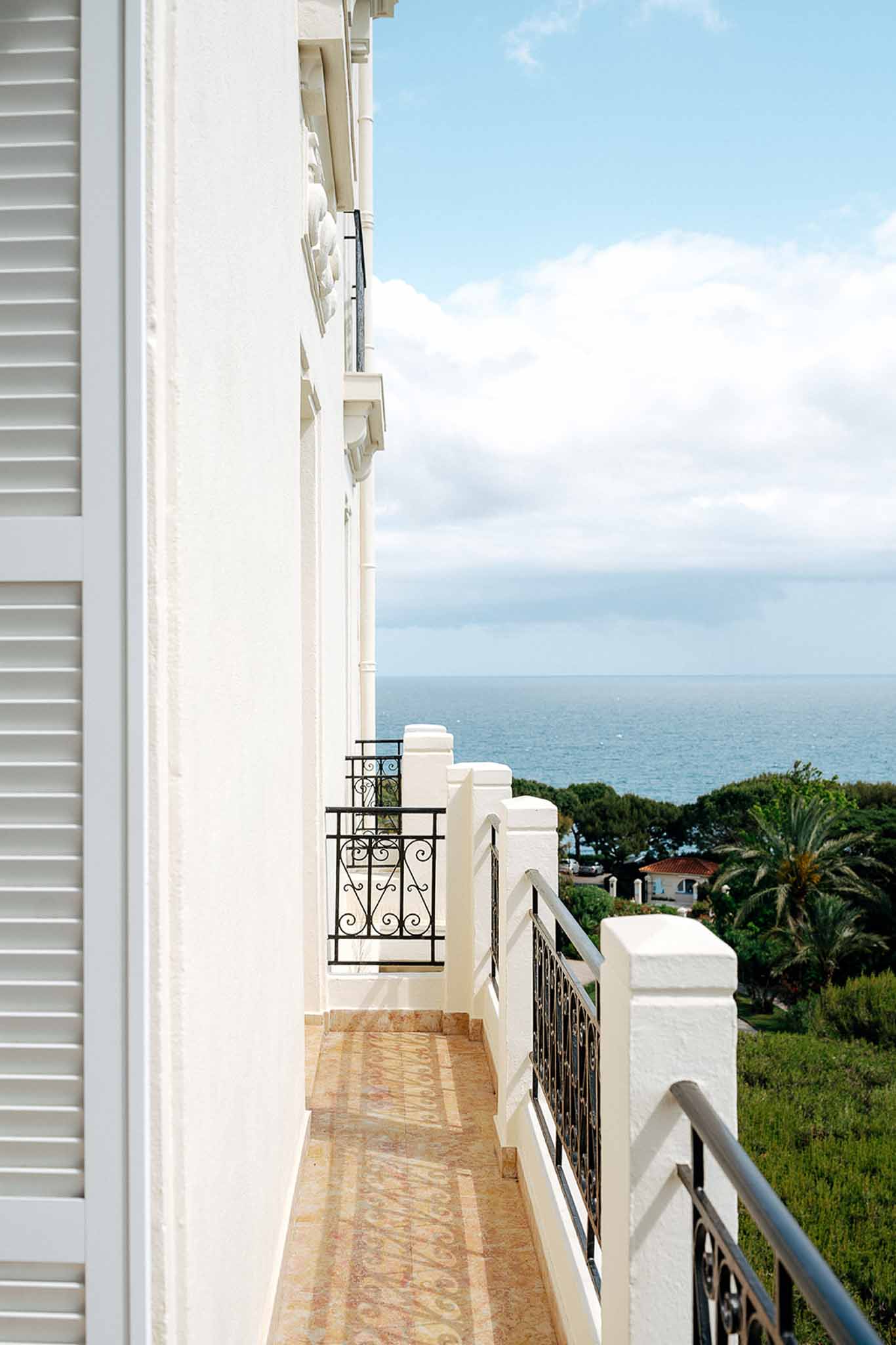 White villa balcony with wrought-iron railings and louvred shutters overlooking palm trees and Mediterranean sea horizon