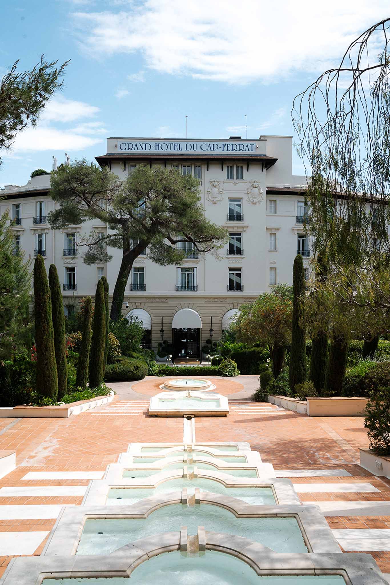 White Belle Epoque hotel with tiered stone fountains, cypress trees, and formal garden approach