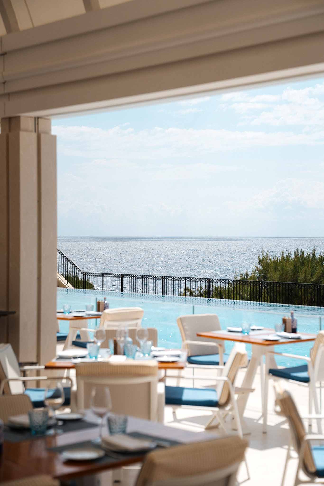 Coastal terrace dining with turquoise glassware, navy cushion chairs, infinity pool, and sea view