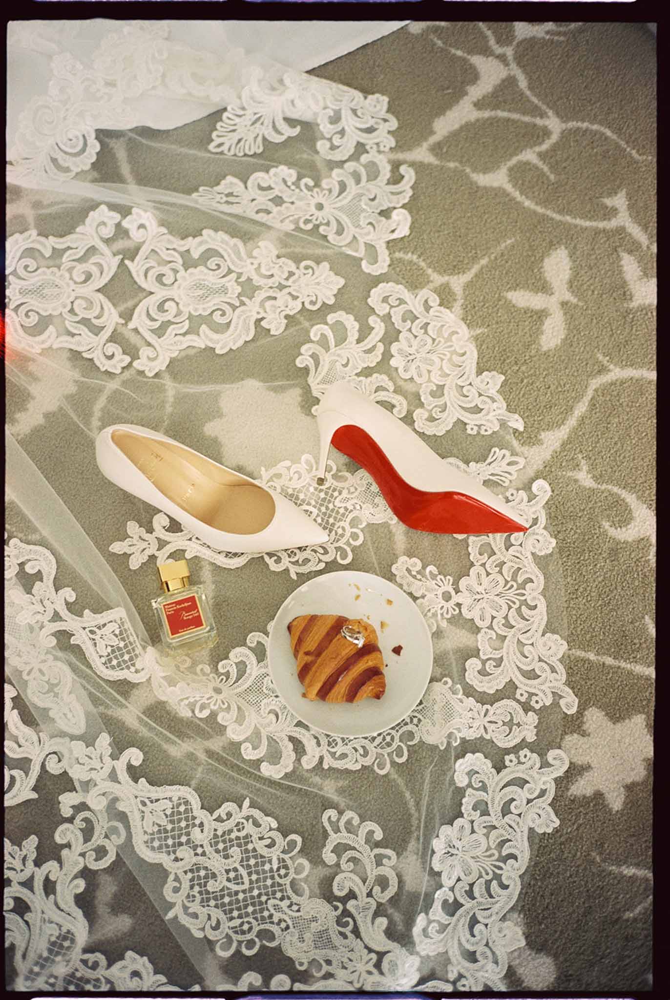 Bridal flat lay with white Louboutin heel, lace veil, perfume bottle, diamond ring and pain au chocolat on grey carpet